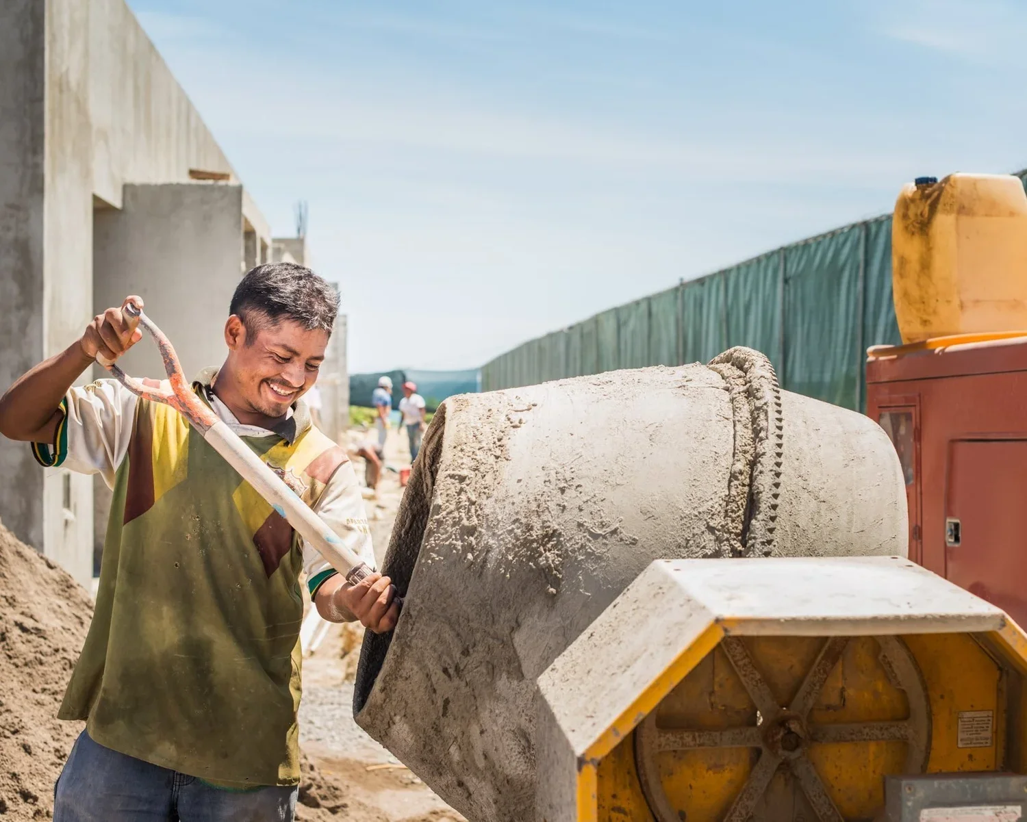 Construction worker smiling as he pours concrete from a cement mixer on a sunny day at a construction site.
