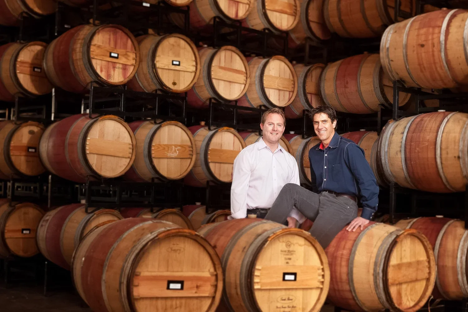 Two men in a wine cellar surrounded by stacked wine barrels, smiling at the camera.