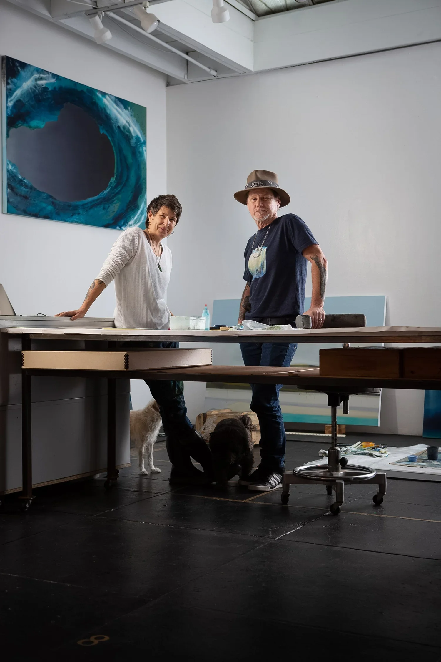 Two artists stand in an art studio with dogs at their feet. One woman and one man, the woman wearing a white sweater and the man wearing a black t-shirt and a wide-brimmed hat, look at the camera. There are paintings and art supplies around them.
