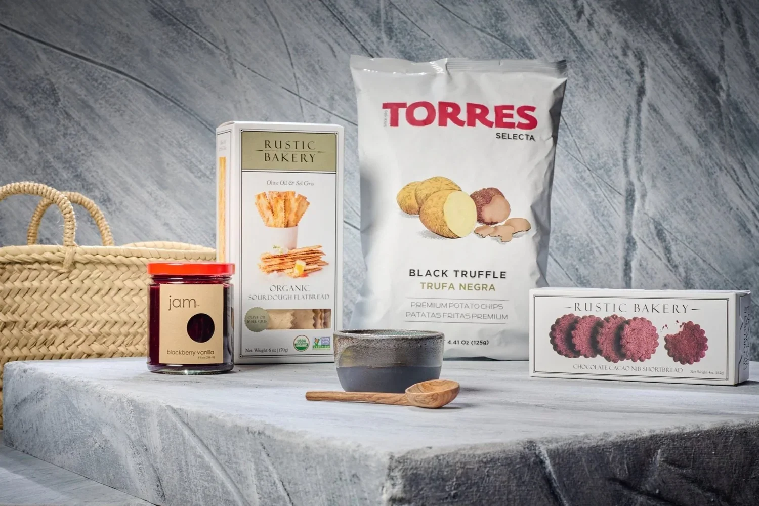 Assorted snacks on a gray marble surface, including a woven basket, a jar of blackberry vanilla jam, a box of organic sourdough flatbreads, a bag of Torres black truffle potato chips, a box of rustic bakery chocolate cacao nib shortbread, and a small