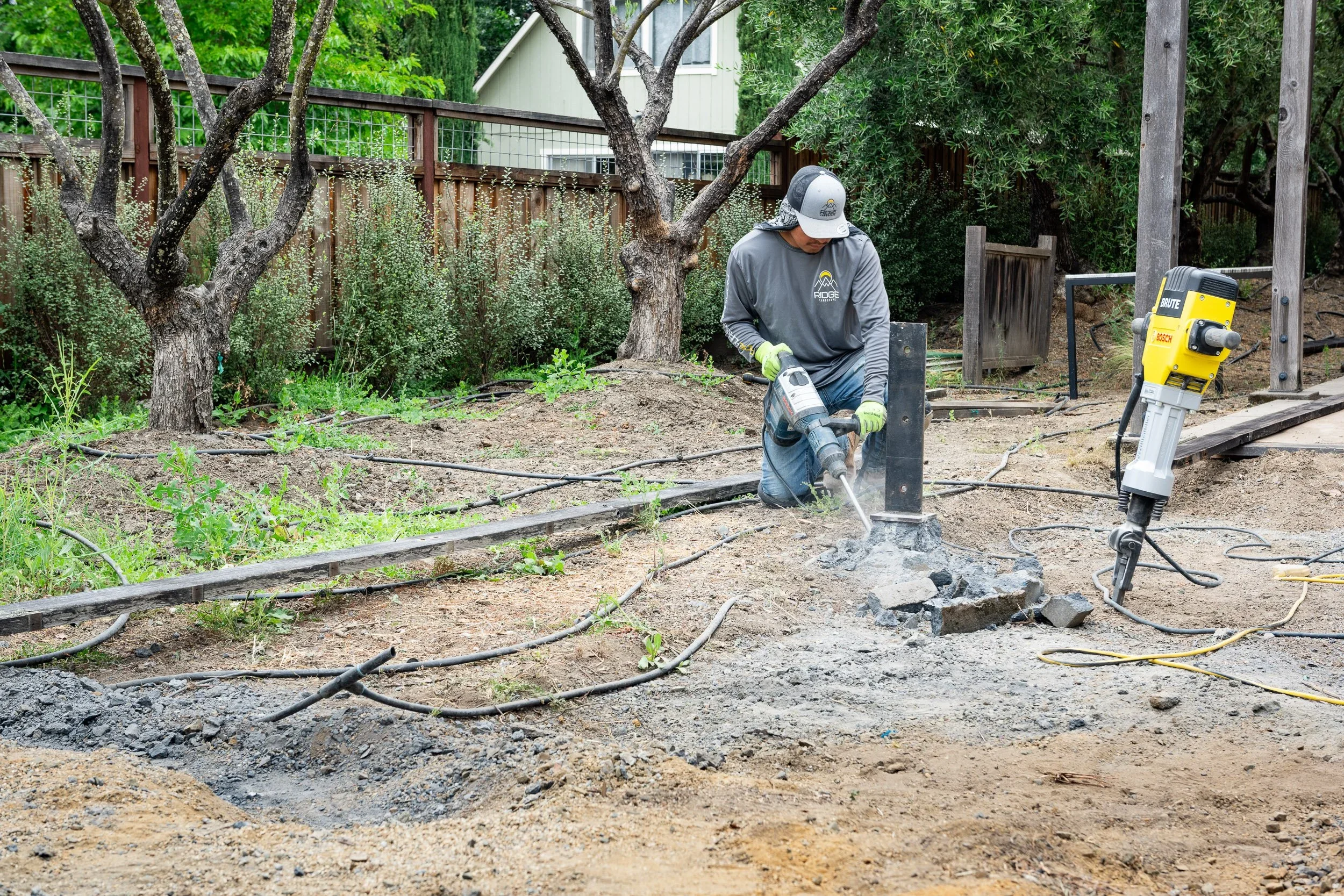 A man kneeling on the ground, using a jackhammer to break concrete in a backyard with trees and a wooden fence.