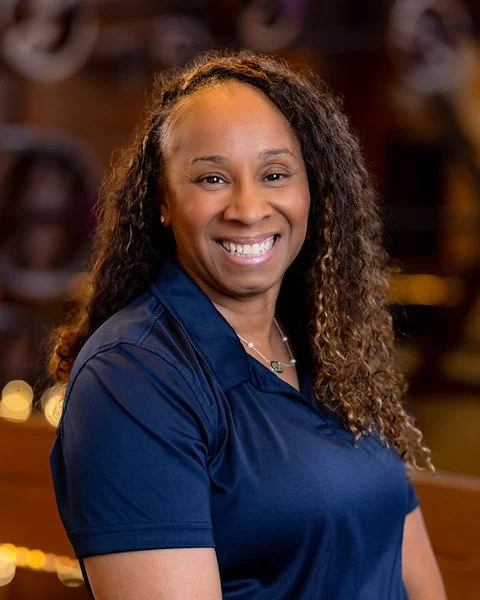 A portrait of a smiling African American woman with shoulder-length hair, wearing a white top, against a dark gray background.