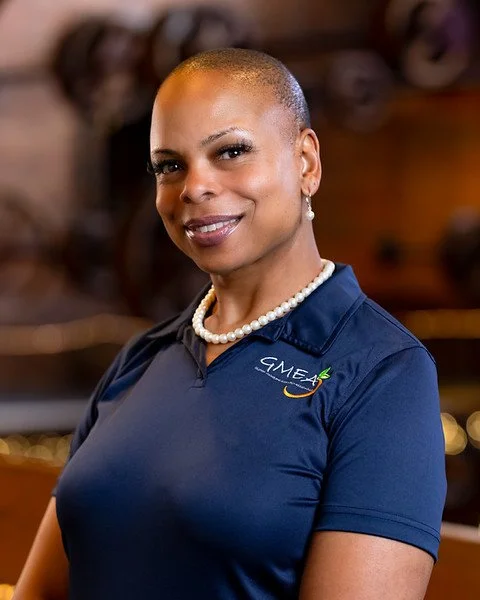 A woman with a shaved head, wearing a black top, hoop earrings, and a watch, smiling with crossed arms in an indoor setting with blurred background.