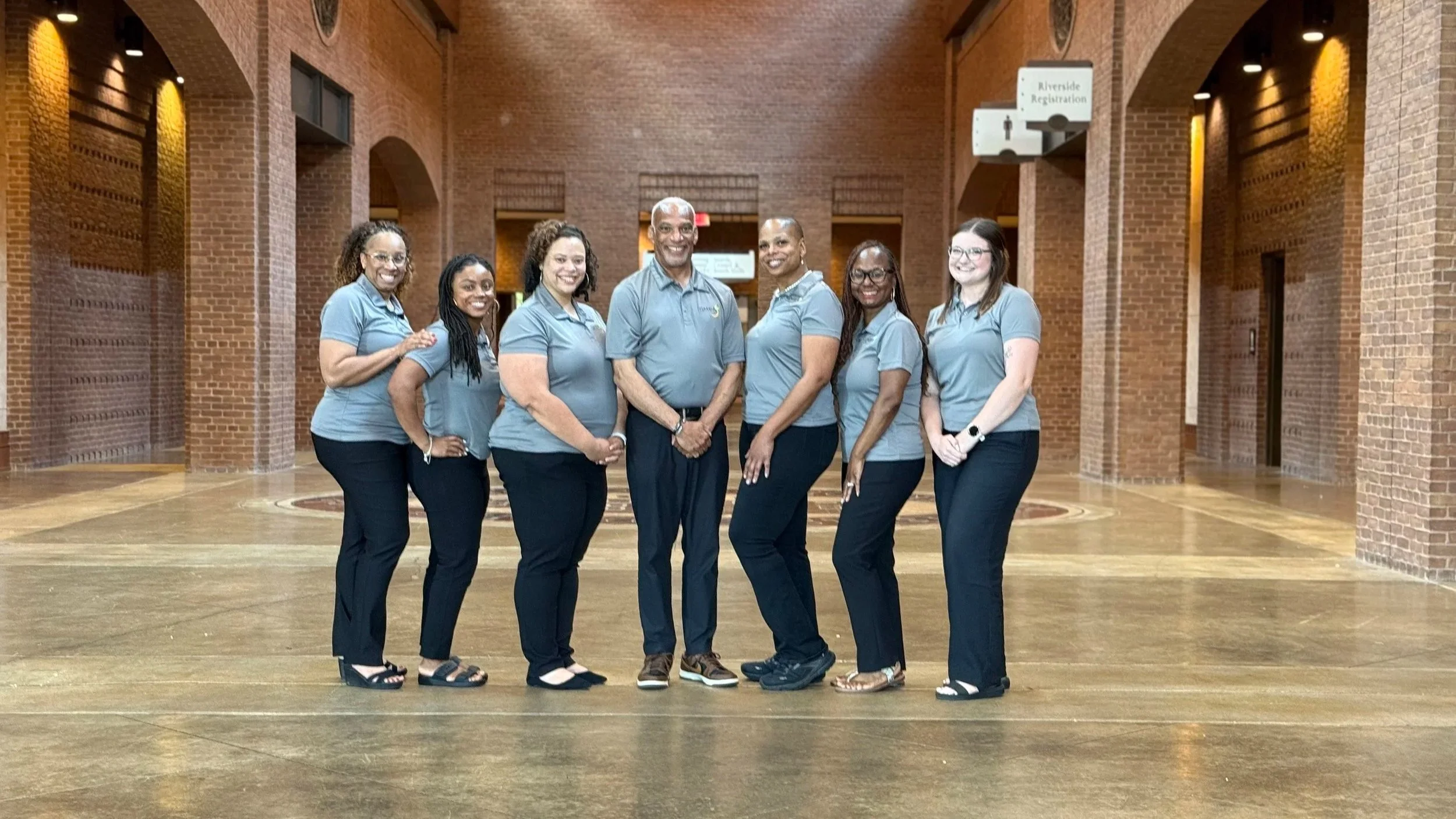 A group of seven diverse people wearing matching light blue shirts and dark pants standing together in a brick-walled indoor space.