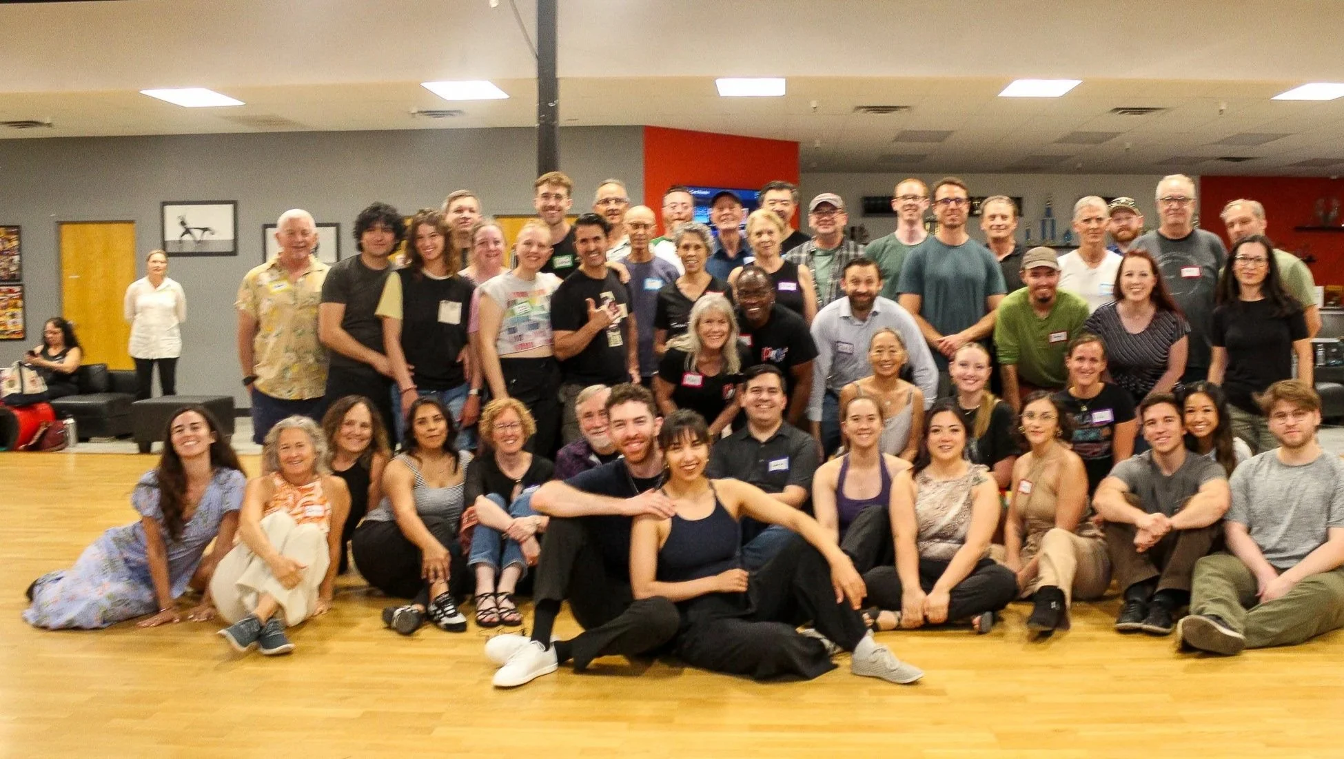 A large group of diverse people gathered indoors, posing for a group photo with some smiling and hugging, in a room with wooden flooring and decorated walls.