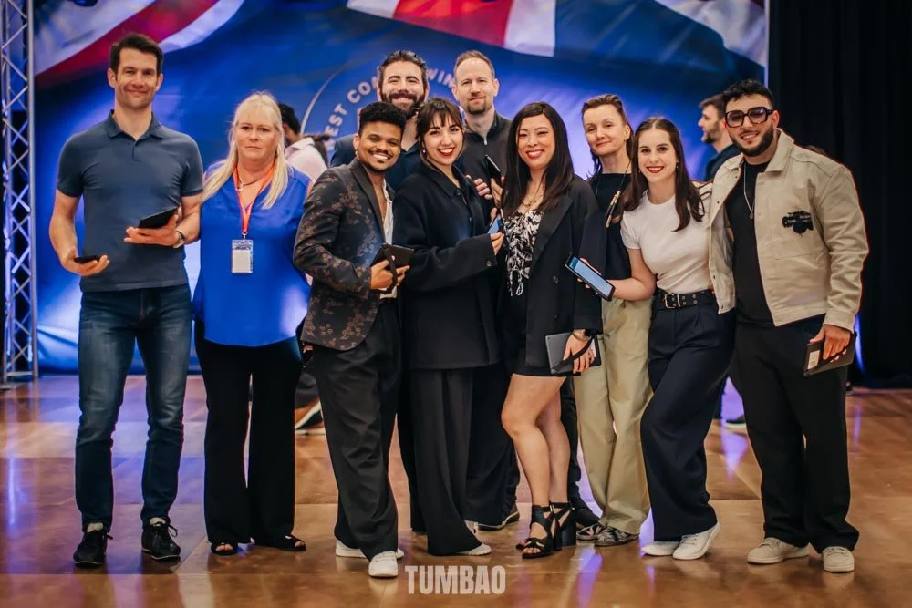 Group of ten people standing together on a stage, posing for a photo at an event with a banner and flags in the background.