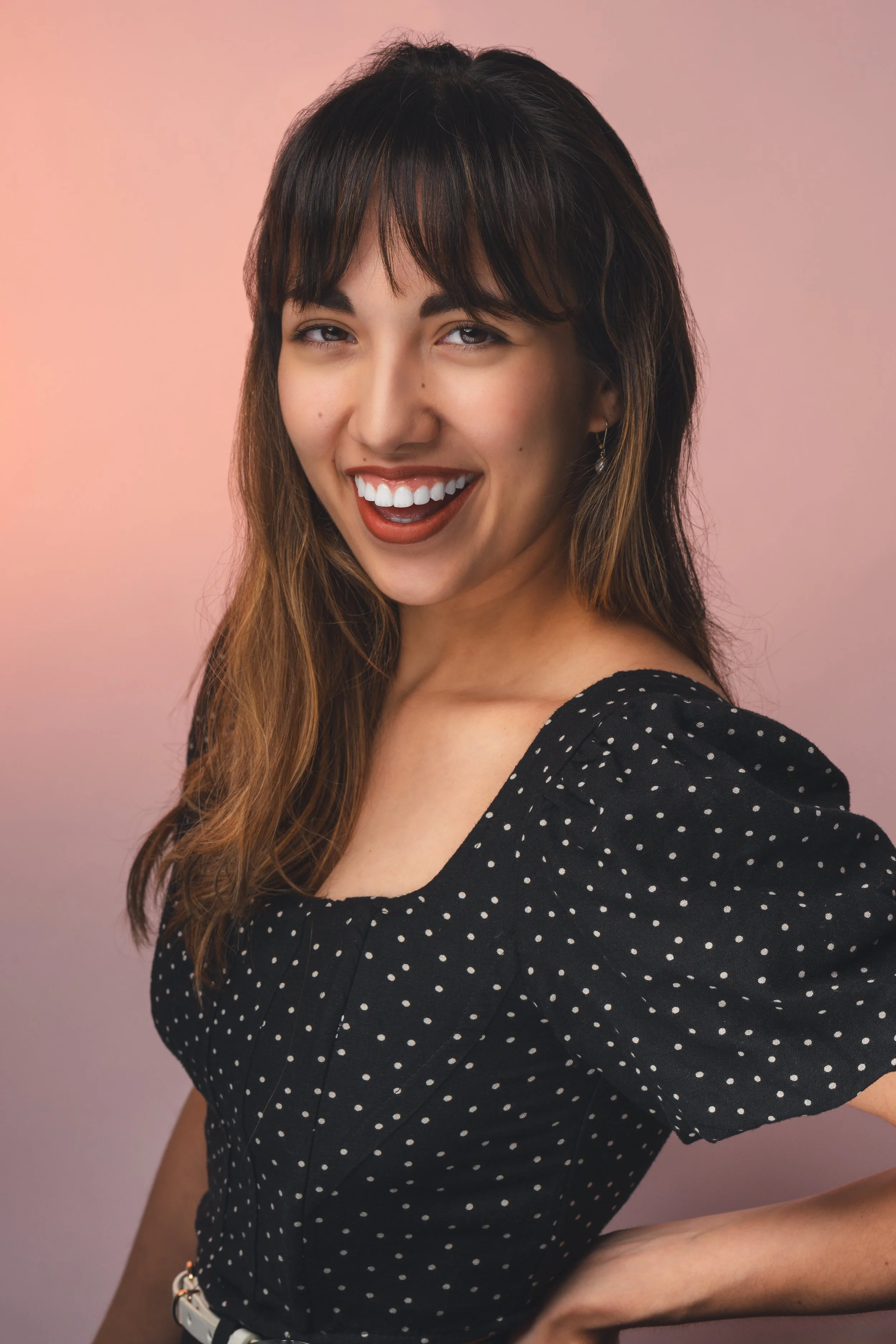 A young woman smiling with red lipstick, wearing a black dress with white polka dots and earrings, standing against a pink gradient background.