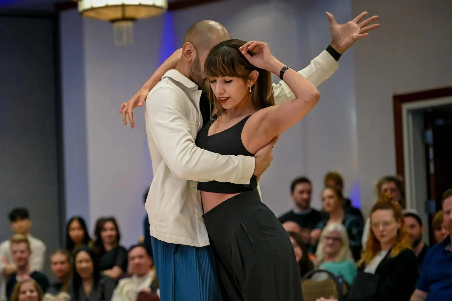 A man and woman are dancing closely at a formal event, with an audience watching in the background.