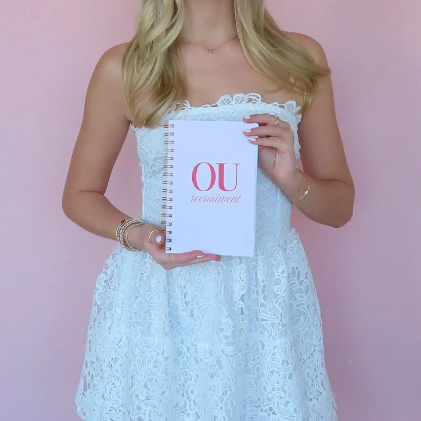 Woman holding a notebook with 'OU recruitment' written on the cover, wearing a white lace dress and standing against a pink background.