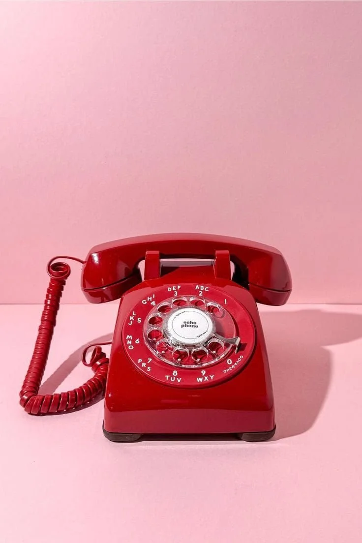A vintage red rotary phone placed on a pink surface against a pink wall background.