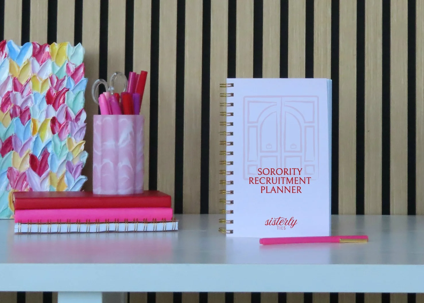 A desk organizer with colorful pens, a notebook titled 'Sorority Recruitment Planner', a pink pen in front, and a decorative paper with pastel colors in the background, set against a wood slat wall.