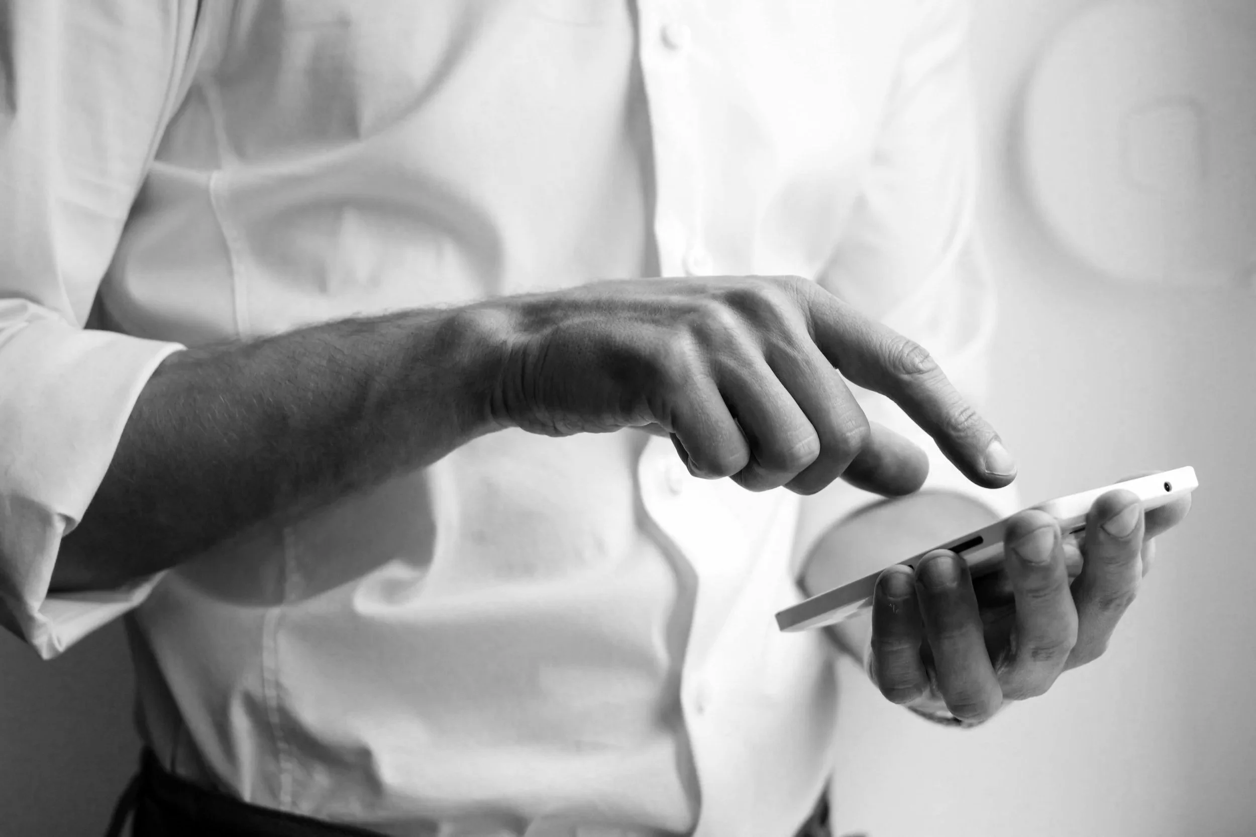 Person holding a smartphone while sitting at a wooden table with a laptop, a remote, and a set of keys on the table.