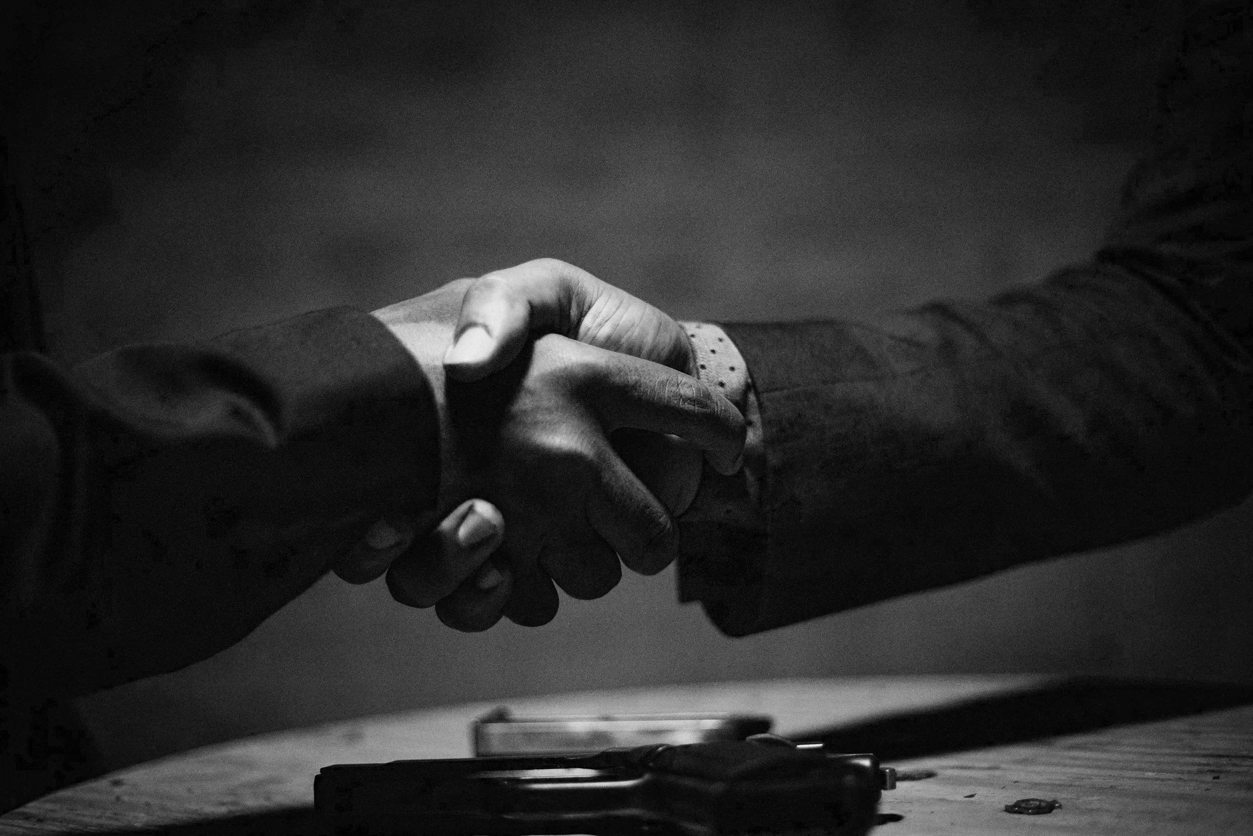 Two people shaking hands over a wooden table, one wearing a dark suit jacket and the other in a black shirt, with a car key and small object on the table.