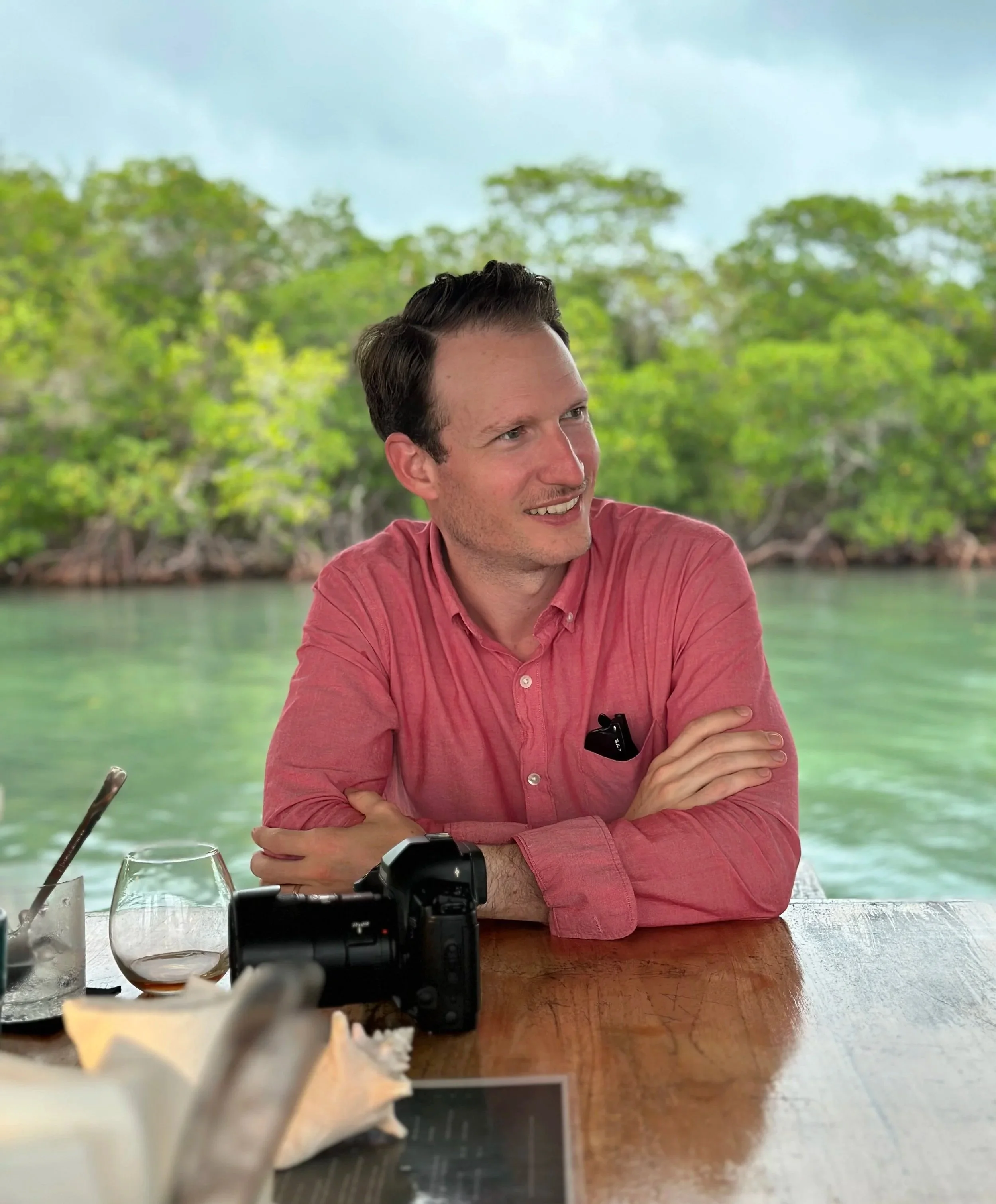 A man with dark hair, wearing a pink shirt, sitting at a wooden table outdoors with a tropical background of green trees and water, smiling and looking to the side.
