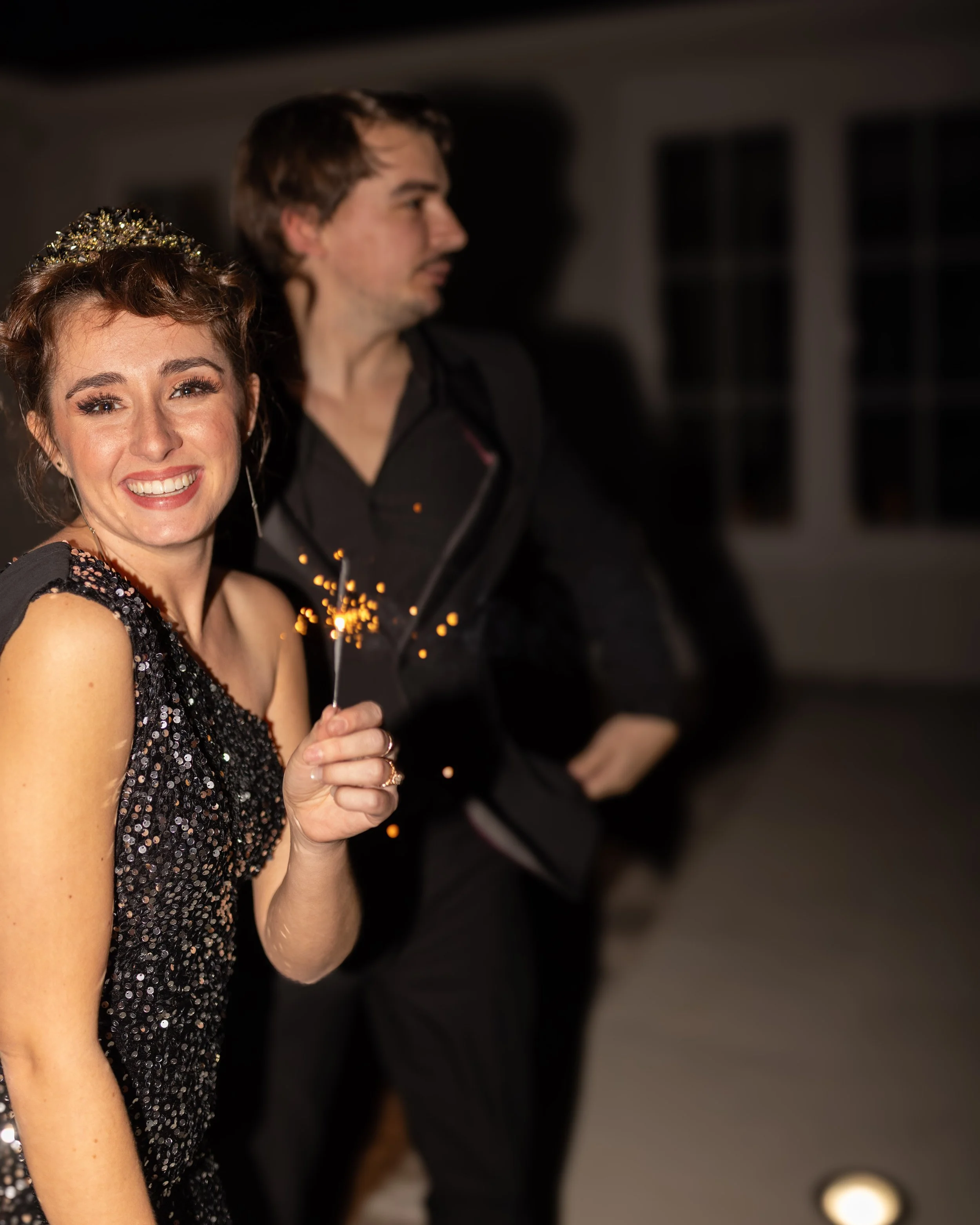A young woman at a celebration holding a sparkler, smiling, dressed in a black sequined dress, with a man in the background at night.