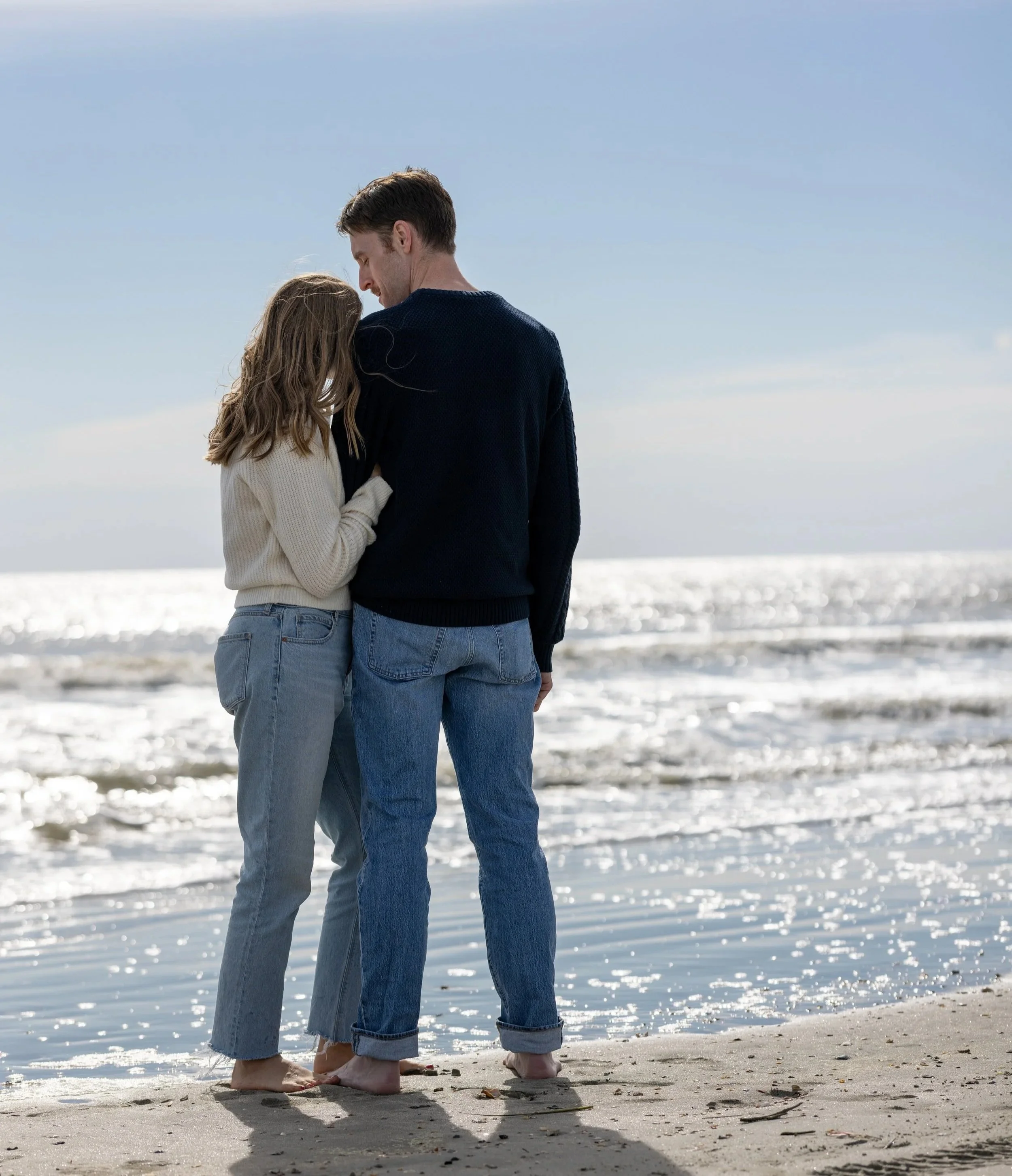 A couple standing close together on the beach, facing each other, with the ocean in the background.