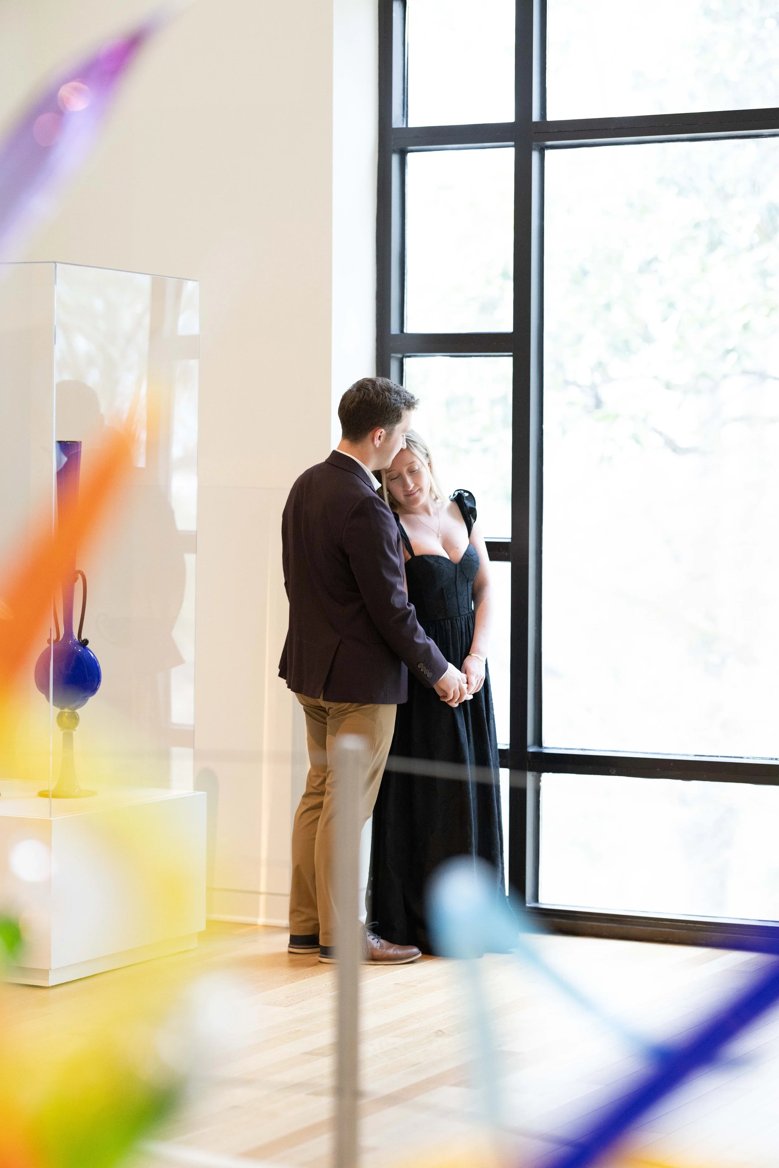 A man and woman stand close together inside a building near a large window, with the man leaning his head against the woman's forehead, both with eyes closed, conveying an intimate moment.