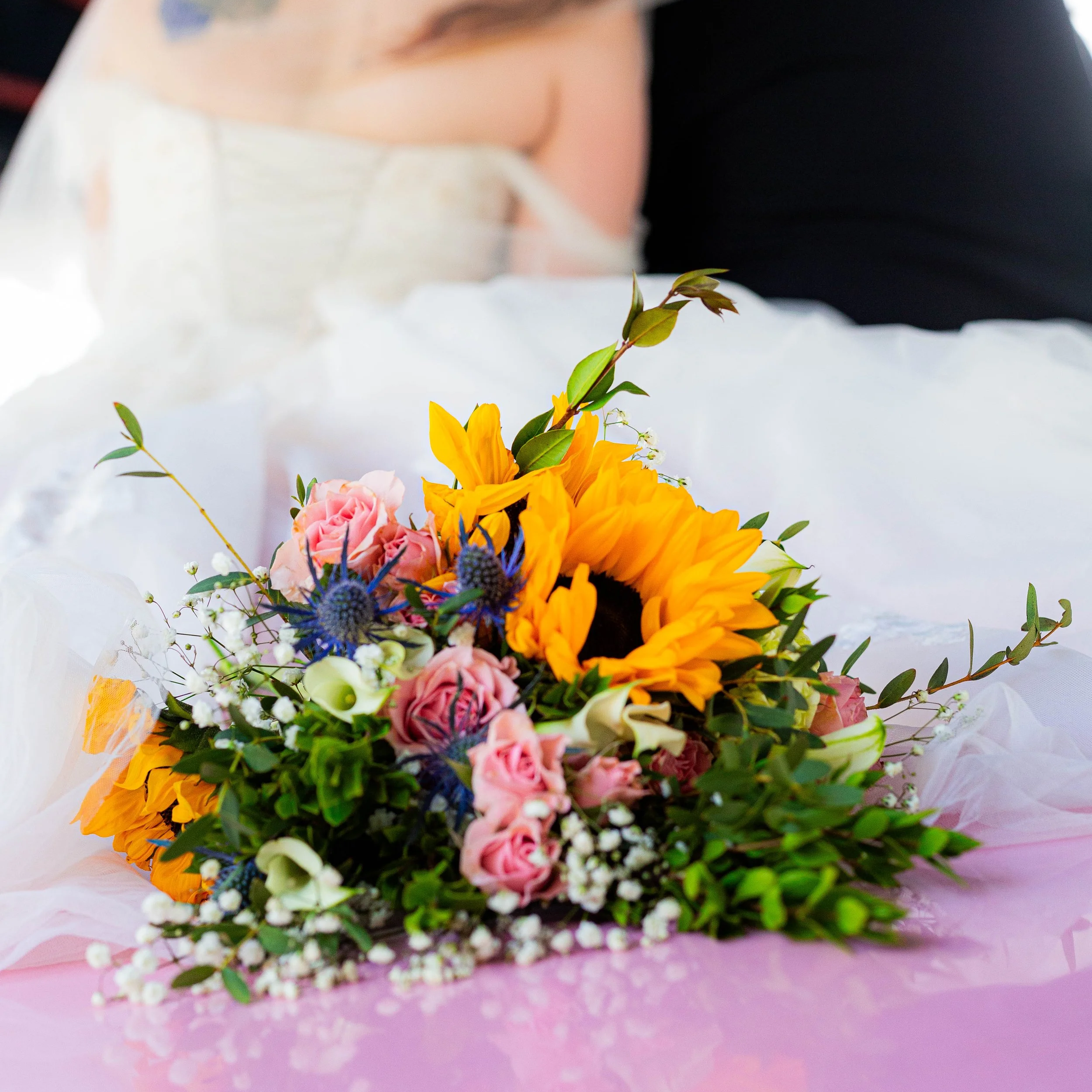 Colorful bouquet of sunflowers, pink roses, white calla lilies, and other flowers resting on the hood of a pink Cadillac car.