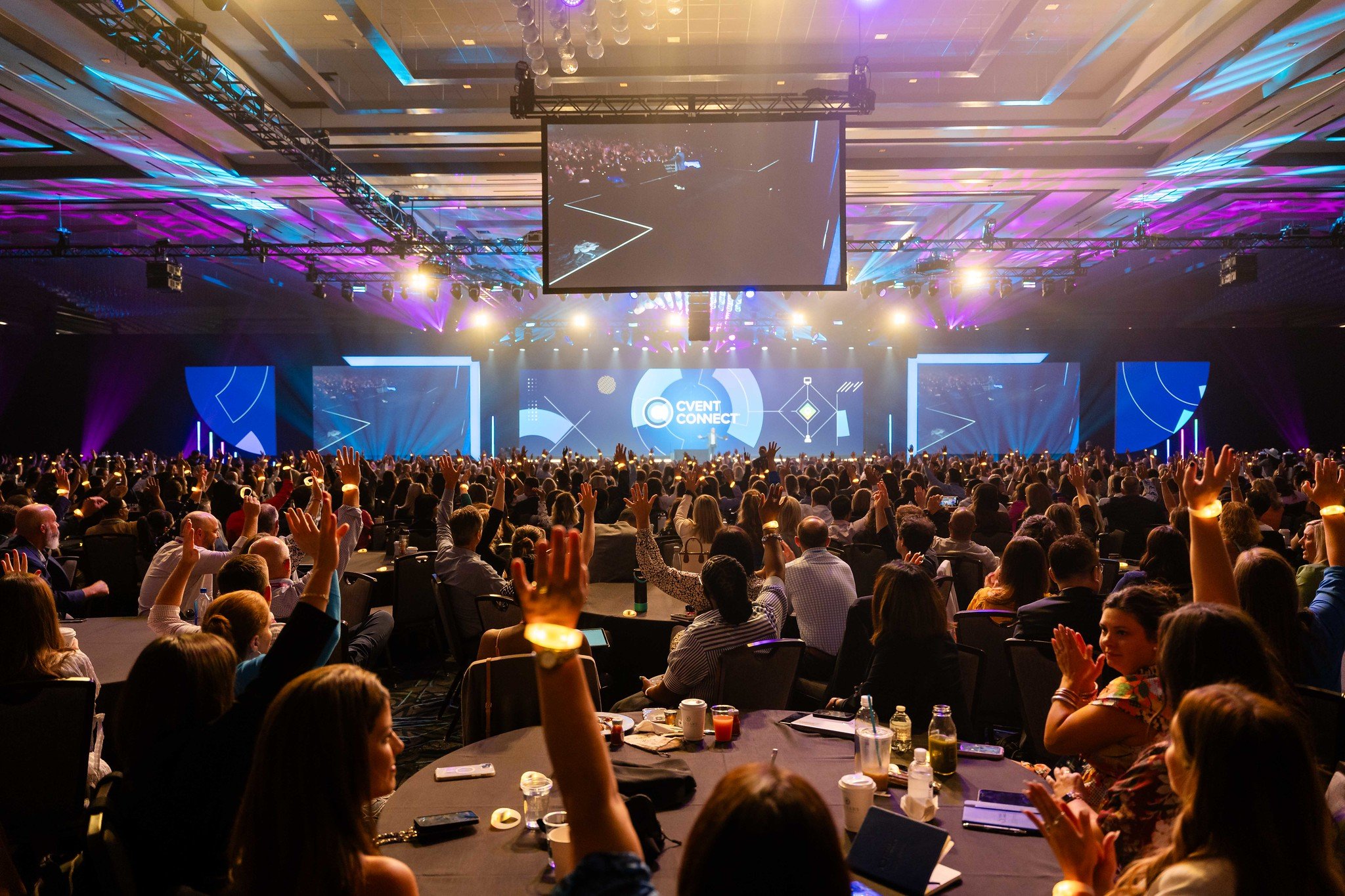 Large conference with many attendees seated at round tables, raising hands towards stage with large screens displaying a presentation, colorful lighting, and a central screen showing the event theme 'Cvent Connect'.