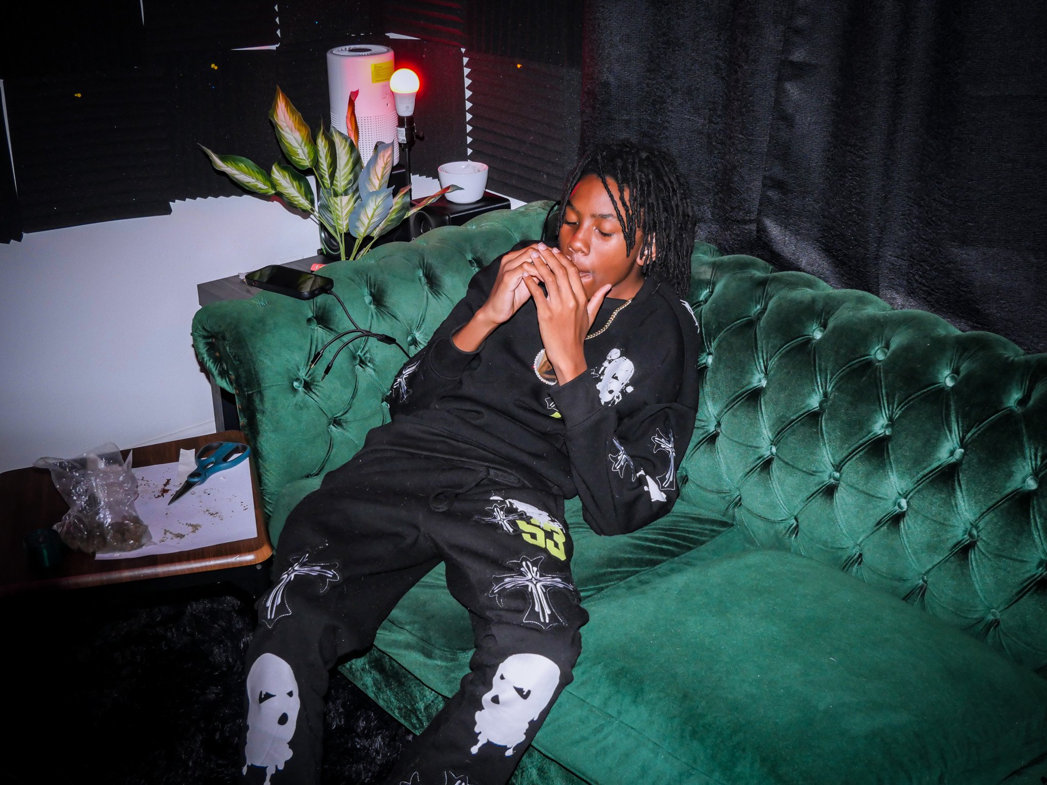 A person with dreadlocks sitting on a green velvet sofa in a dimly lit room, wearing black clothing with skull and face designs, holding a cigarette, with a side table holding a pair of scissors, some paper, and a plastic bag, a potted plant, a lamp,