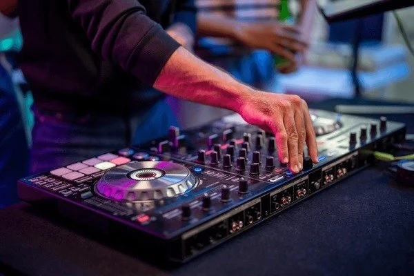Close-up of a person adjusting controls on a DJ mixer, with a turntable and other DJ equipment visible.