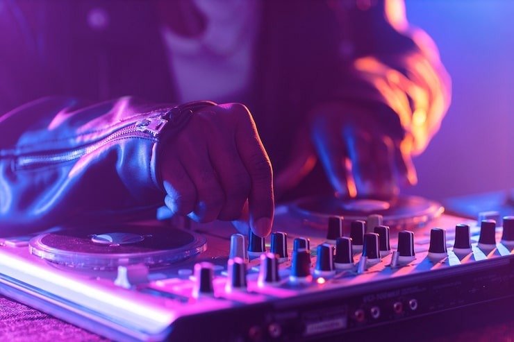 A DJ wearing a leather jacket adjusting controls on a mixer in a dimly lit setting with colorful lighting.