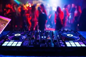 A DJ's mixing console and turntables at a lively nightclub with colorful lights and a dancing crowd in the background.