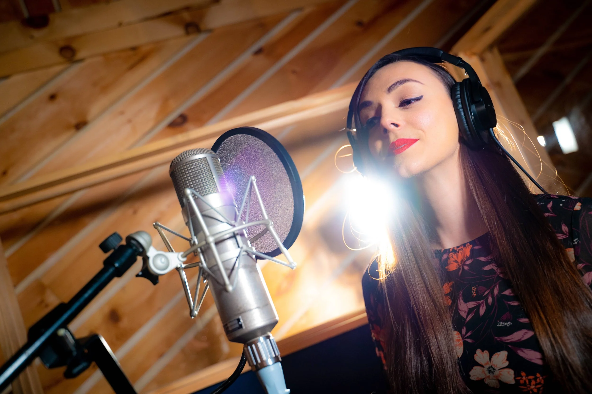 A woman in a recording studio, singing into a microphone with headphones on, with a wooden ceiling and a bright light behind her.