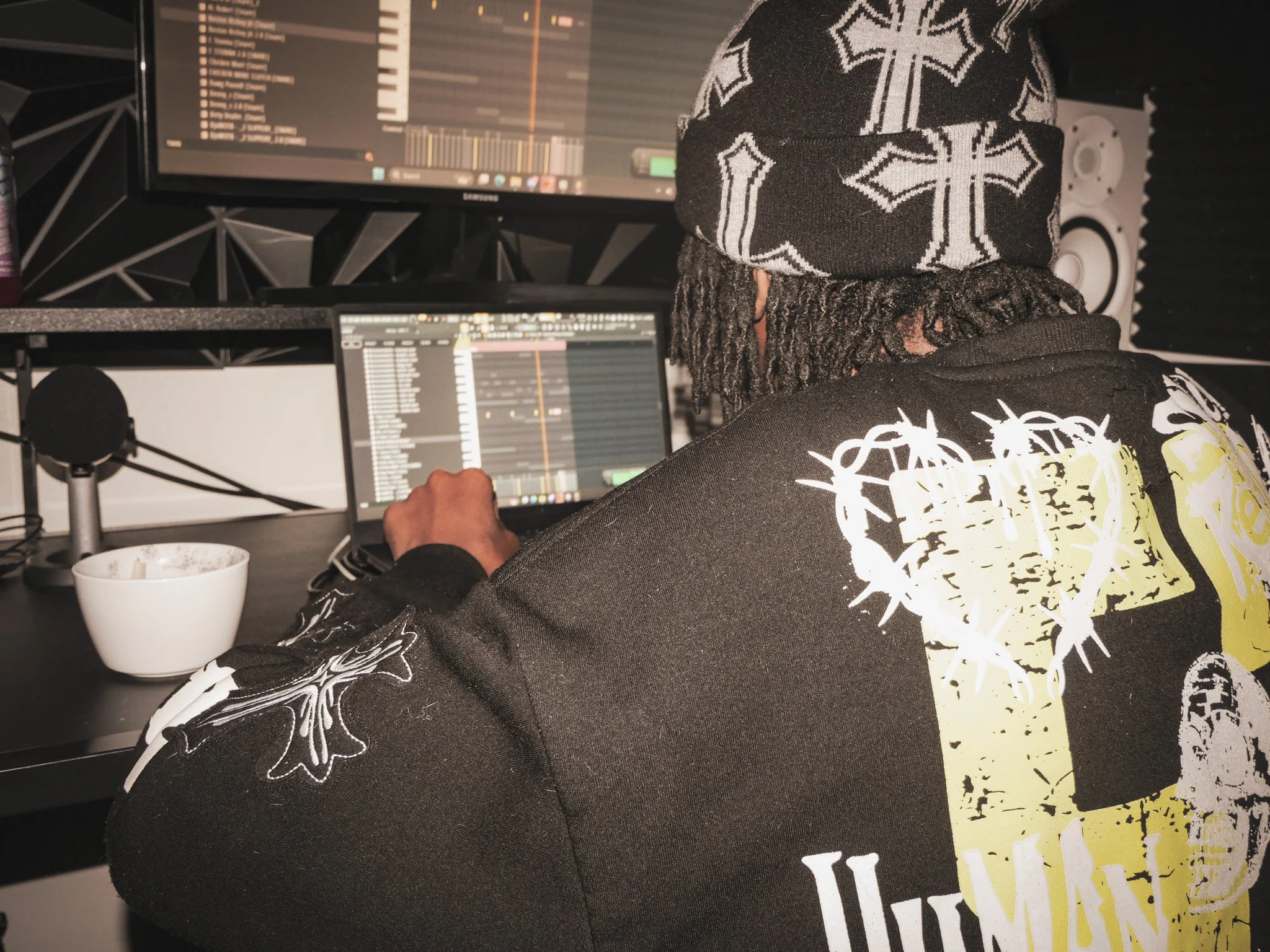 Person working on music production in a studio, wearing a black hoodie with white graphic design, a black beanie with cross patterns, and headphones, with computer monitors displaying music software.