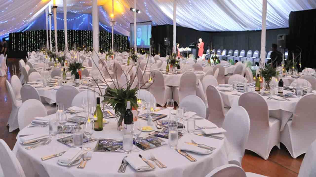 Elegant banquet hall decorated for a formal event with round tables draped in white tablecloths, floral centerpieces, and chairs covered in white slipcovers. There is a stage with a speaker and a large projection screen, and string lights adorn the c