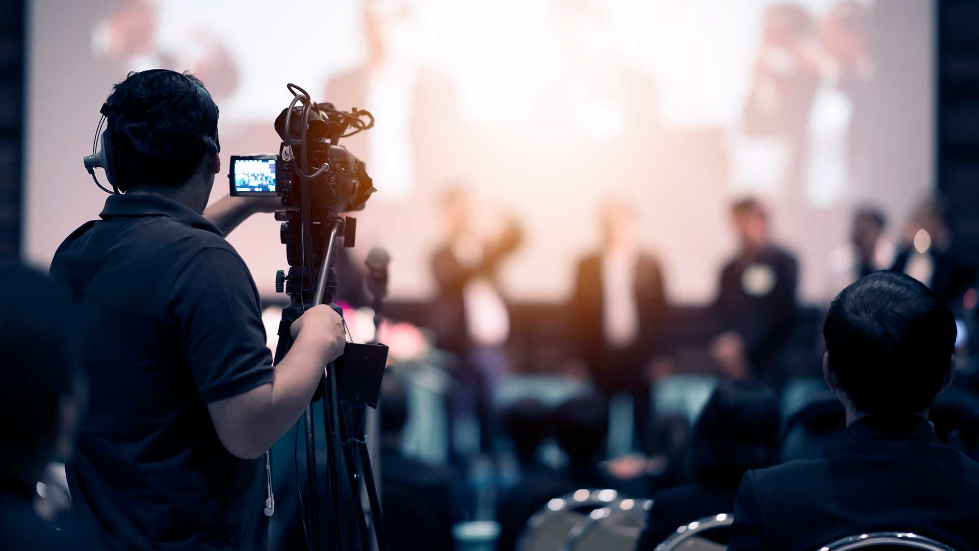 A camera operator recording a panel discussion at a conference. The scene shows a speaker on stage with a blurred background.