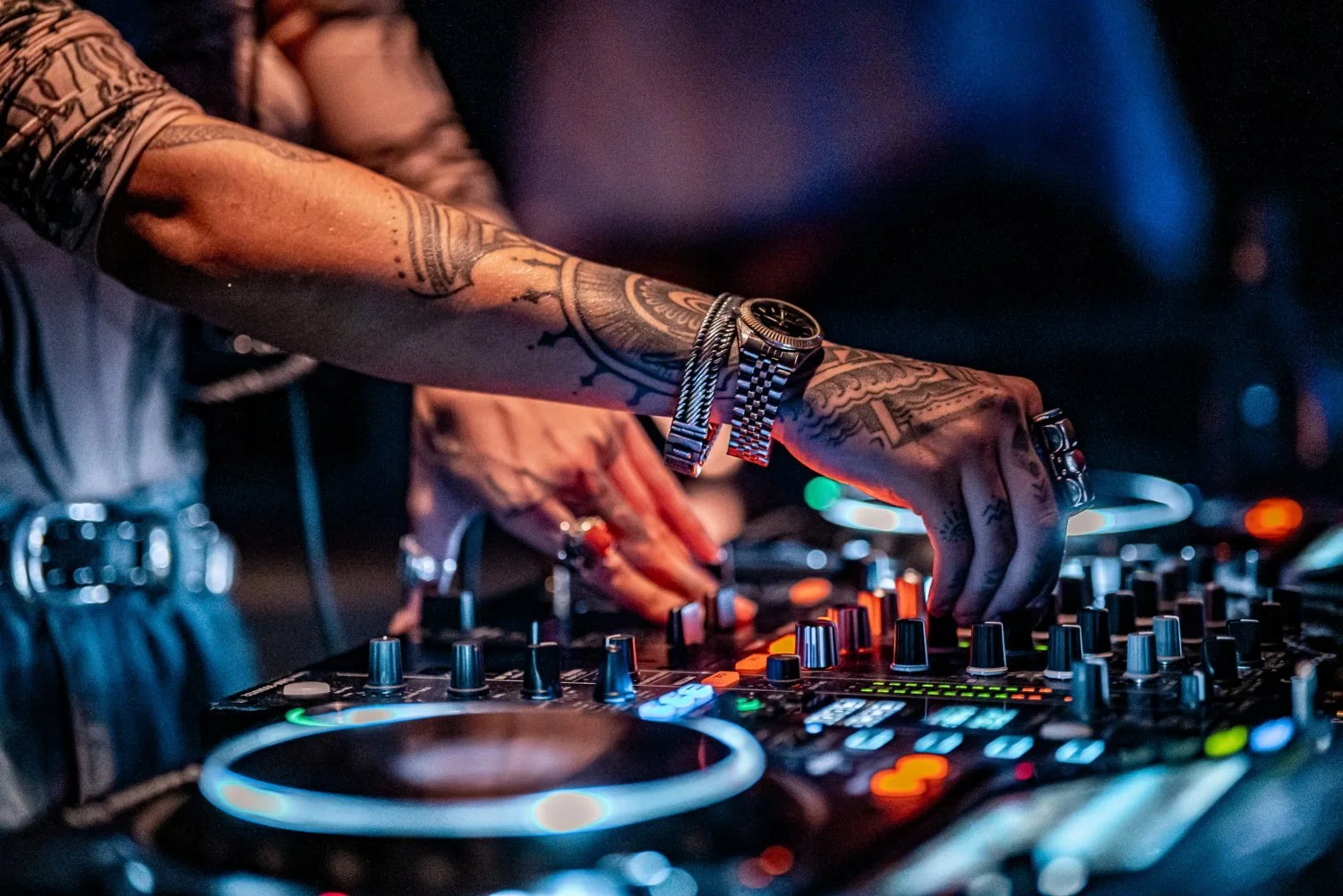 Close-up of a DJ with tattooed arms operating a DJ mixer in a dimly lit environment with colorful lights.