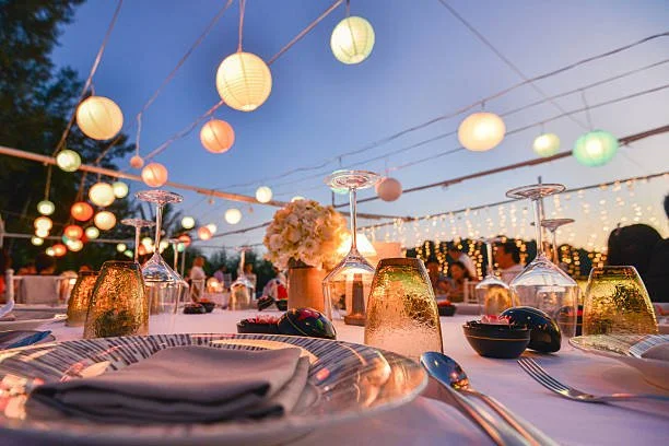 Outdoor dinner setting with hanging paper lanterns and string lights during dusk, with tableware, glasses, and a floral centerpiece.