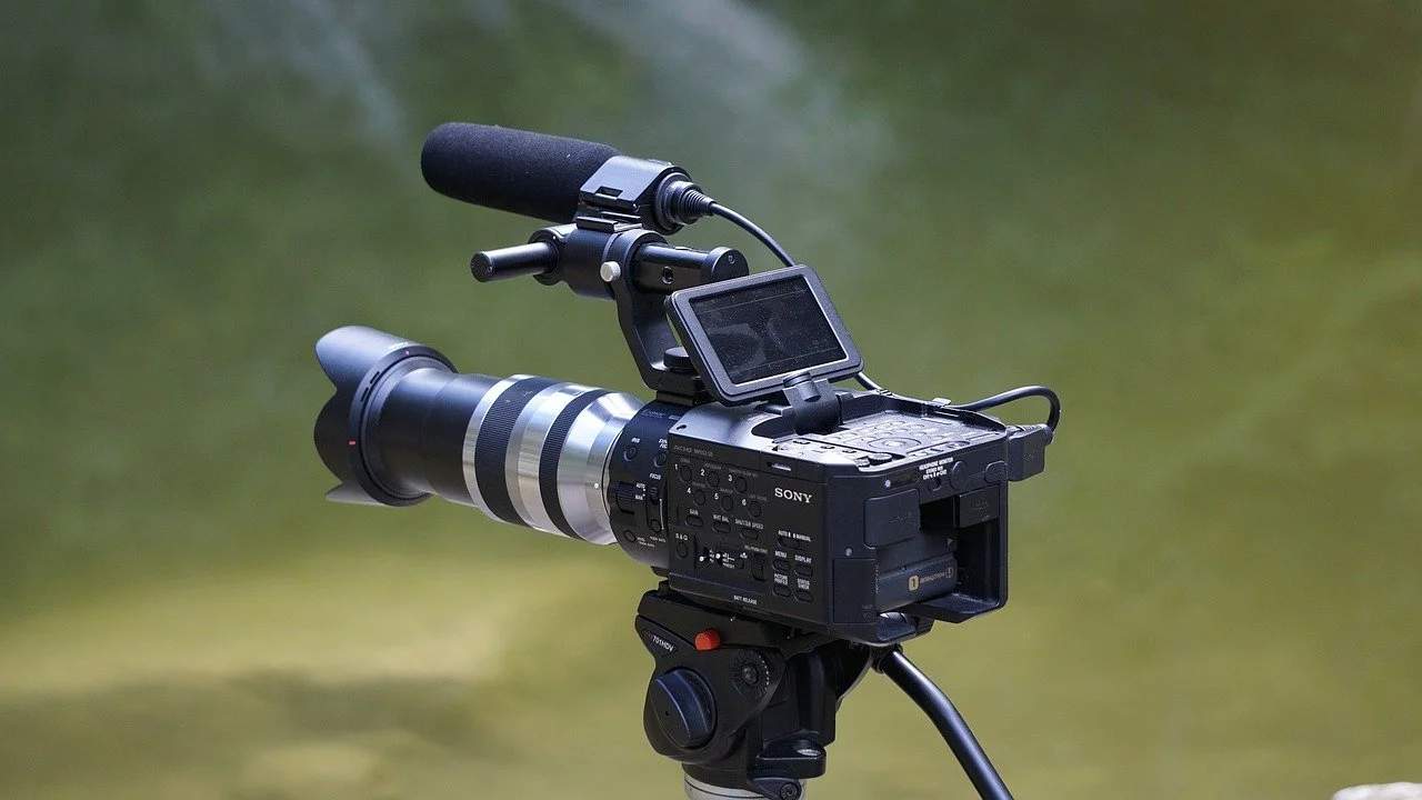 Professional video camera with a microphone mounted on top, set outdoors against a blurred natural background.