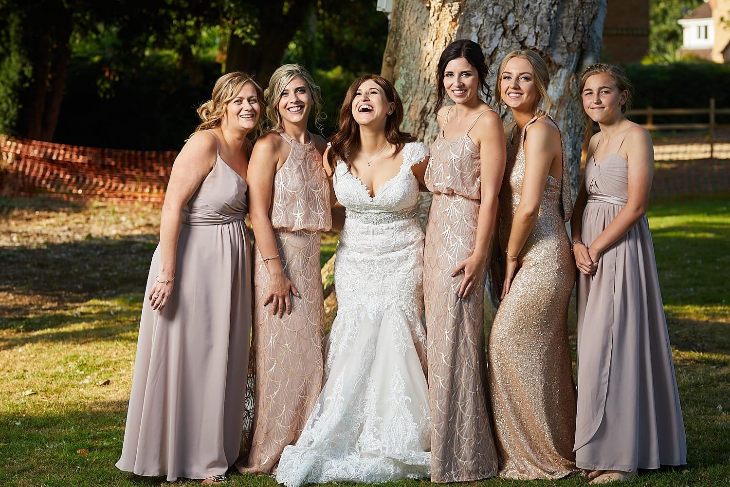 A group of seven women, including a bride in a white wedding gown, standing outdoors in front of a large tree and smiling.