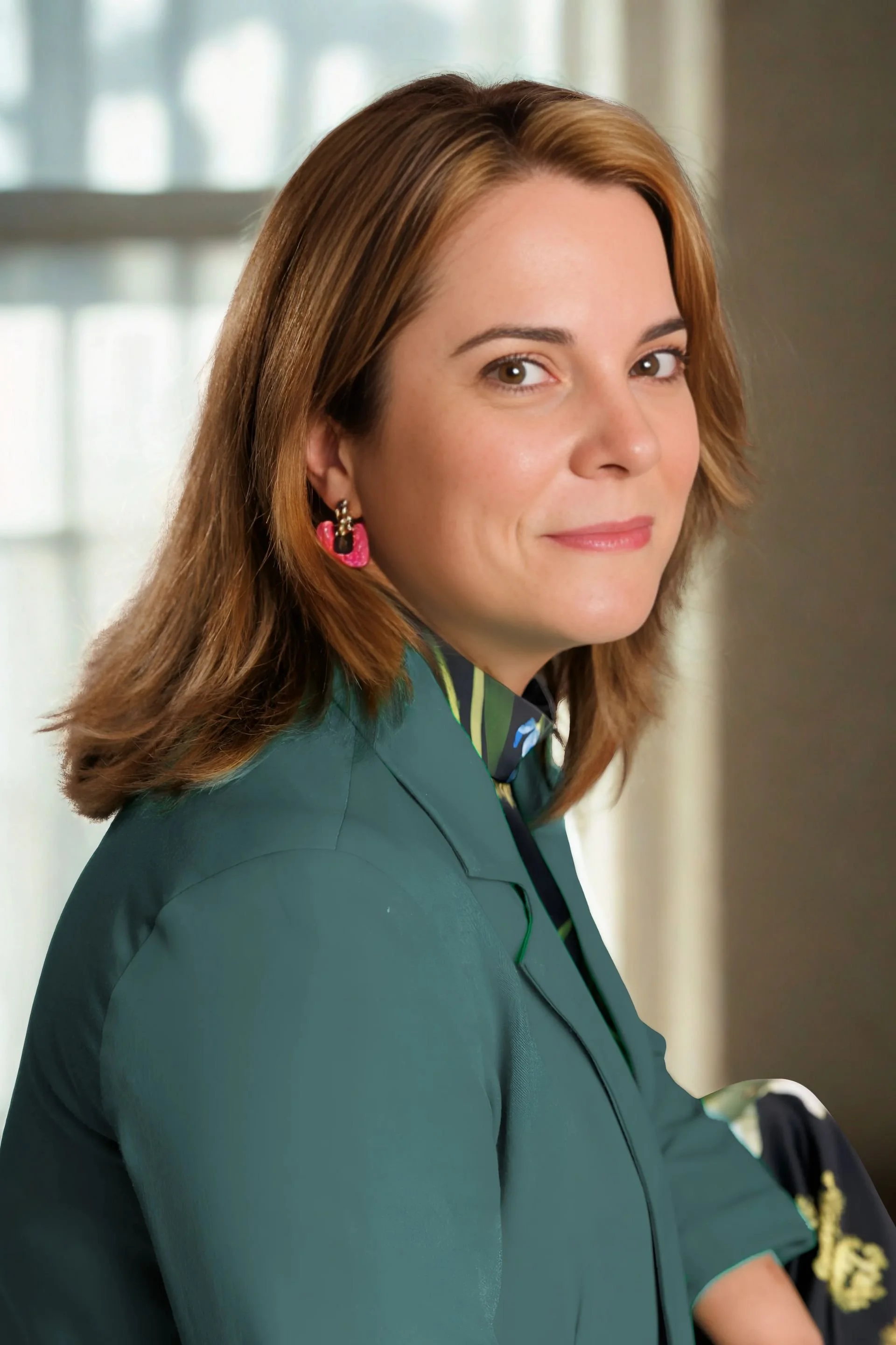rebeca-from-her wealthy club-with shoulder-length light brown hair, wearing a green blazer and a colorful scarf, sitting against a plain light grey background.
