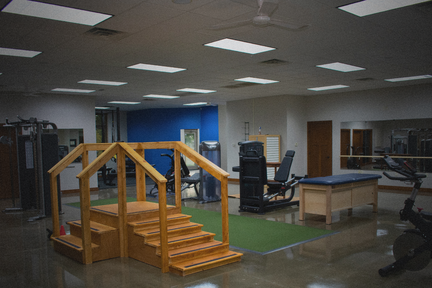 Empty fitness or physical therapy gym with exercise equipment, a small wooden staircase, and mirrors on the wall.