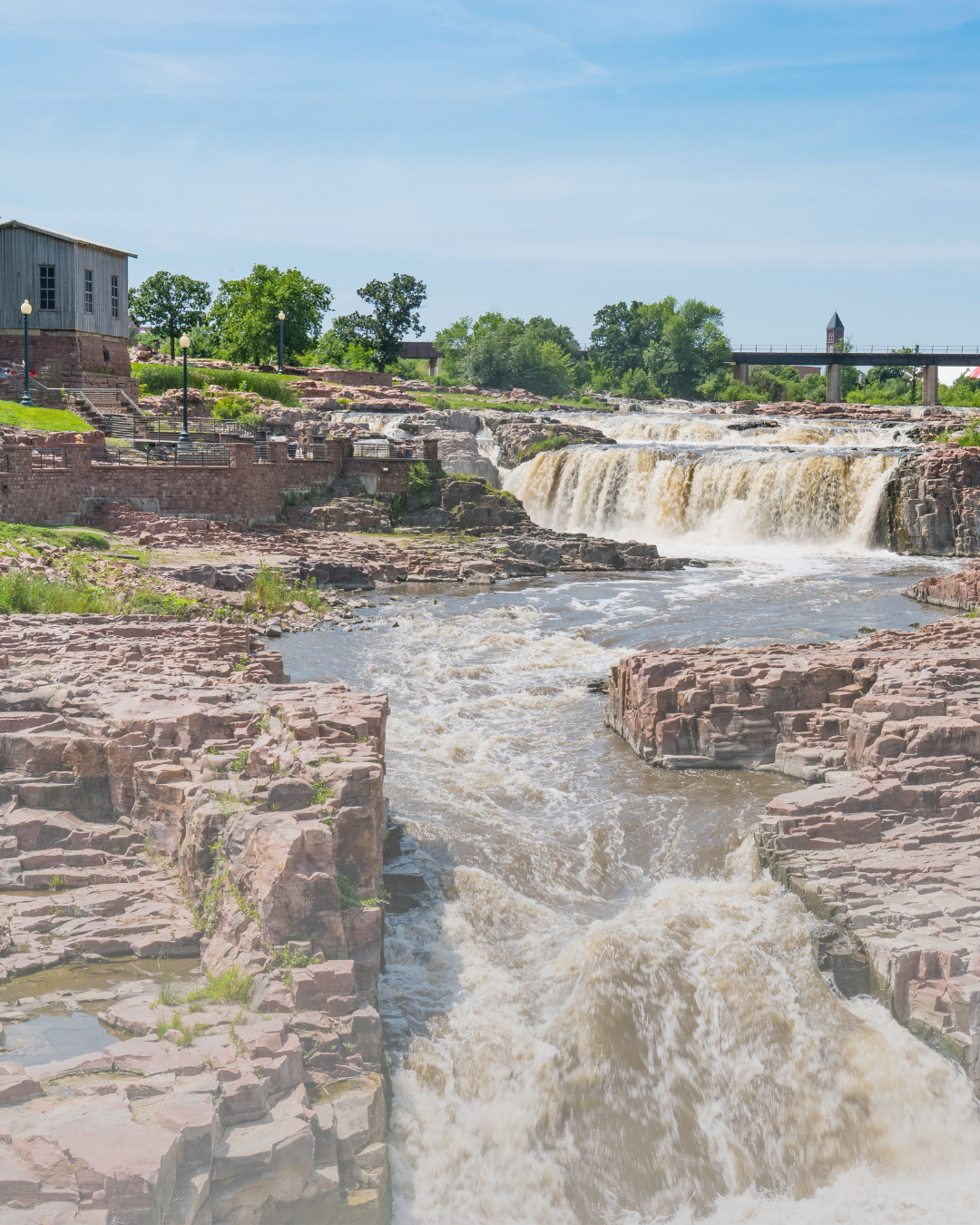 A scenic view of a waterfall with rocky edges, a bridge in the background, and a small building on the left side under a clear blue sky with some clouds.
