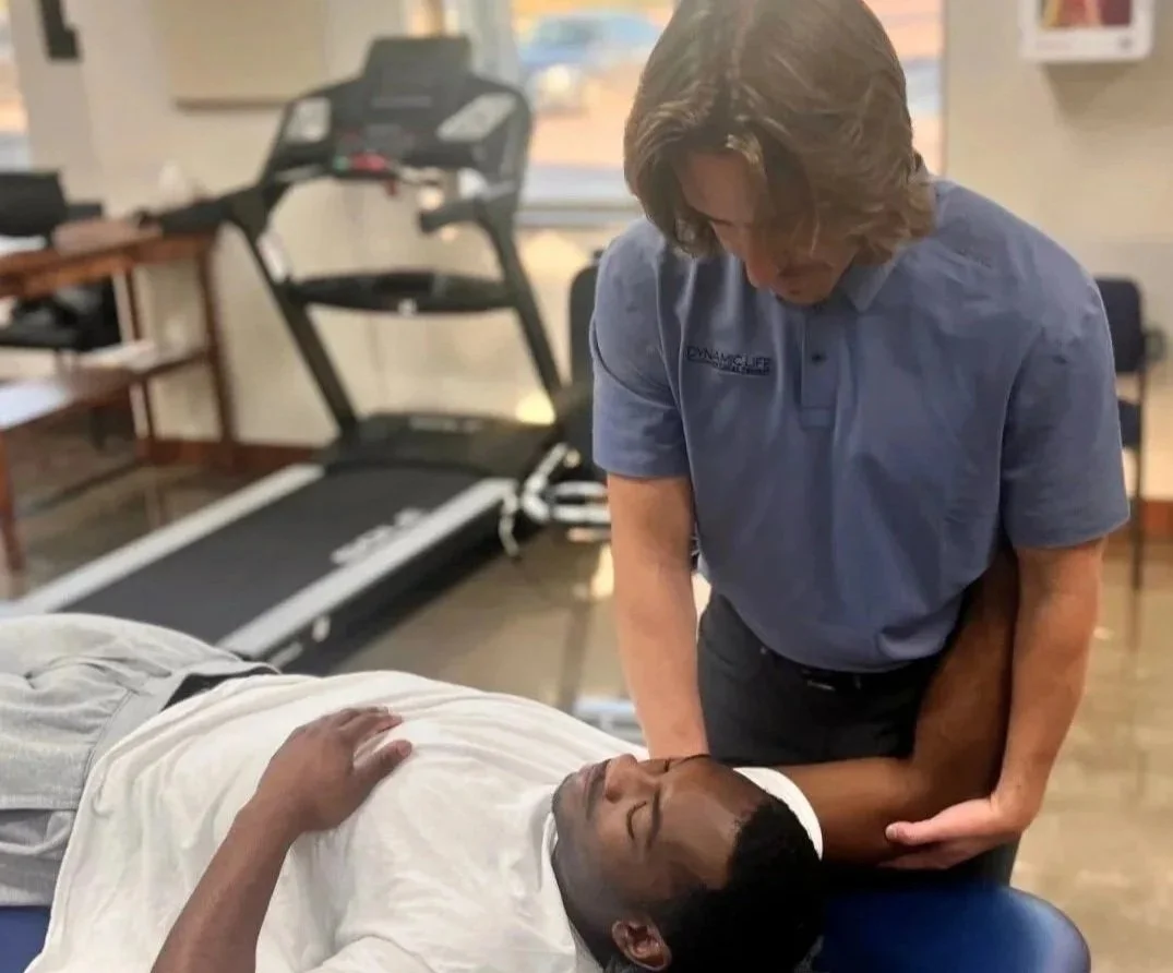 A man receiving physical therapy treatment on his shoulder from a male healthcare professional in a clinical setting with exercise equipment visible in the background.
