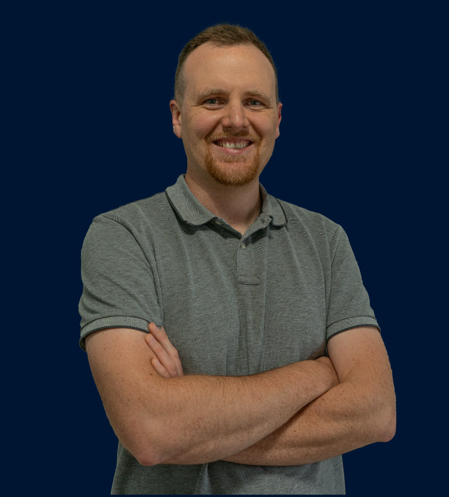A man with short hair and a goatee, wearing a gray polo shirt, is smiling and standing with his arms crossed against a dark blue background.