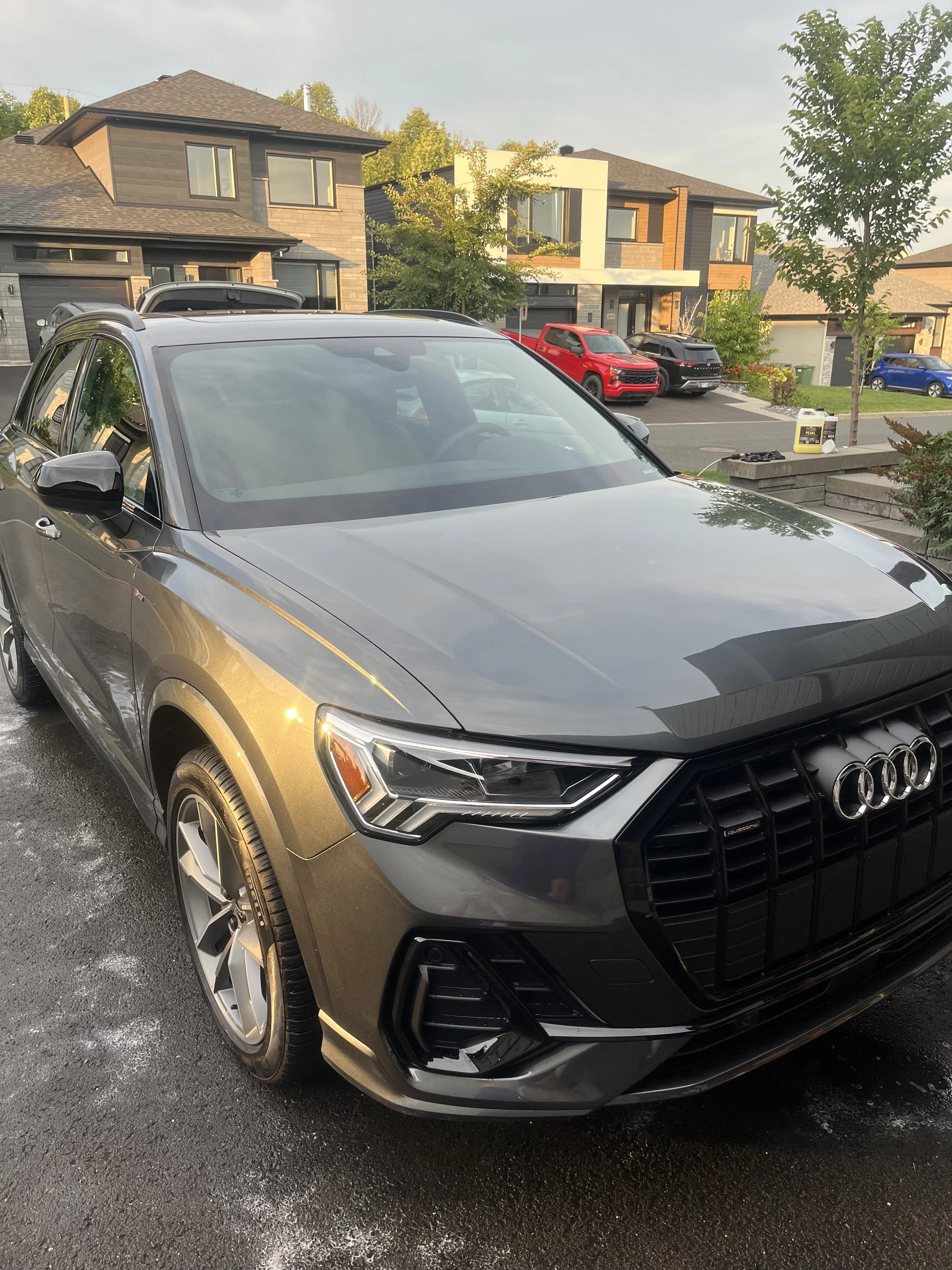 Une voiture Audi grise stationnée devant des maisons modernes dans une rue résidentielle, avec plusieurs autres voitures en arrière-plan.
