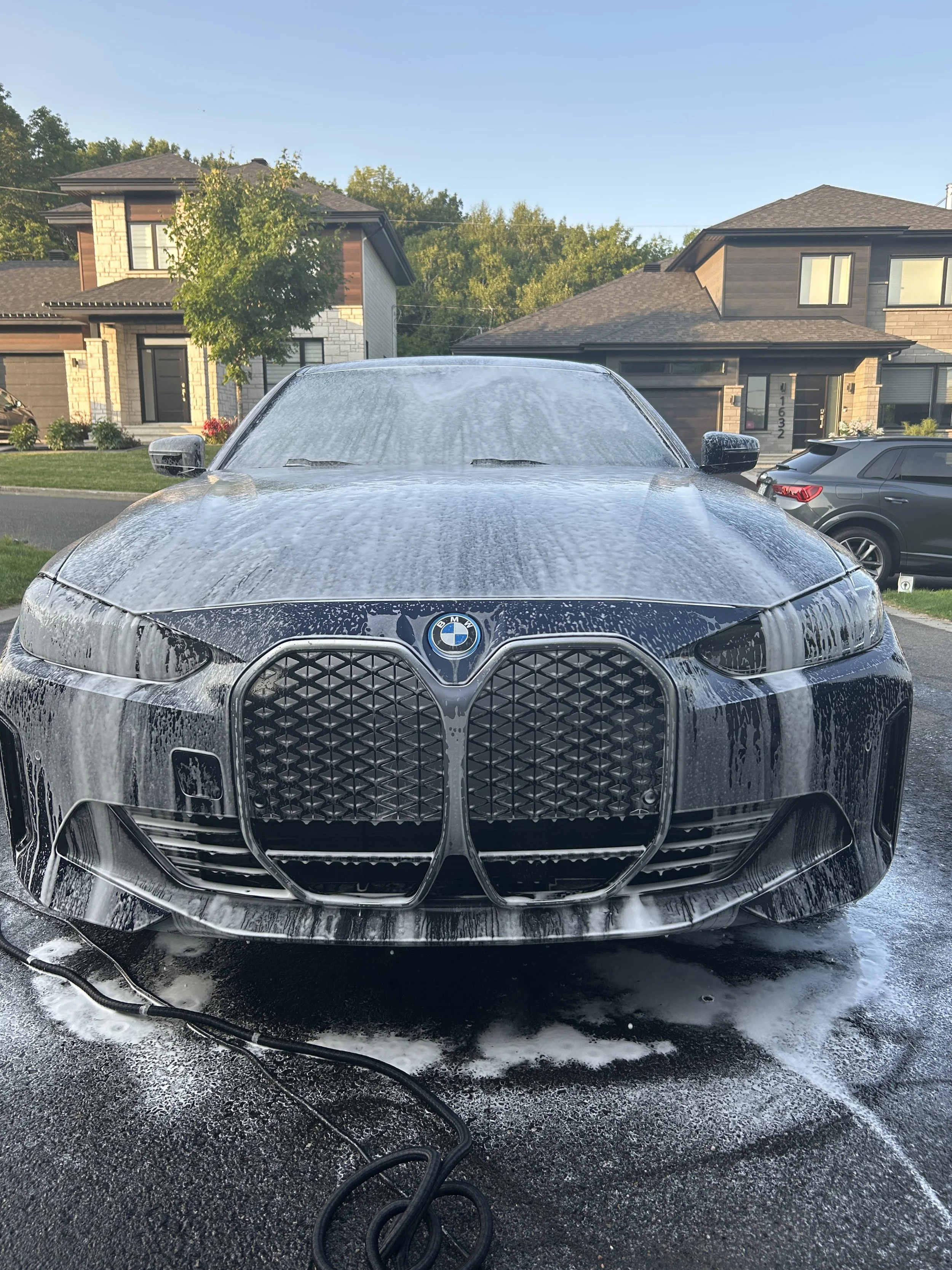 Voiture BMW en train d'être lavée, chevauchée de mousse et de savon, dans une allée résidentielle.