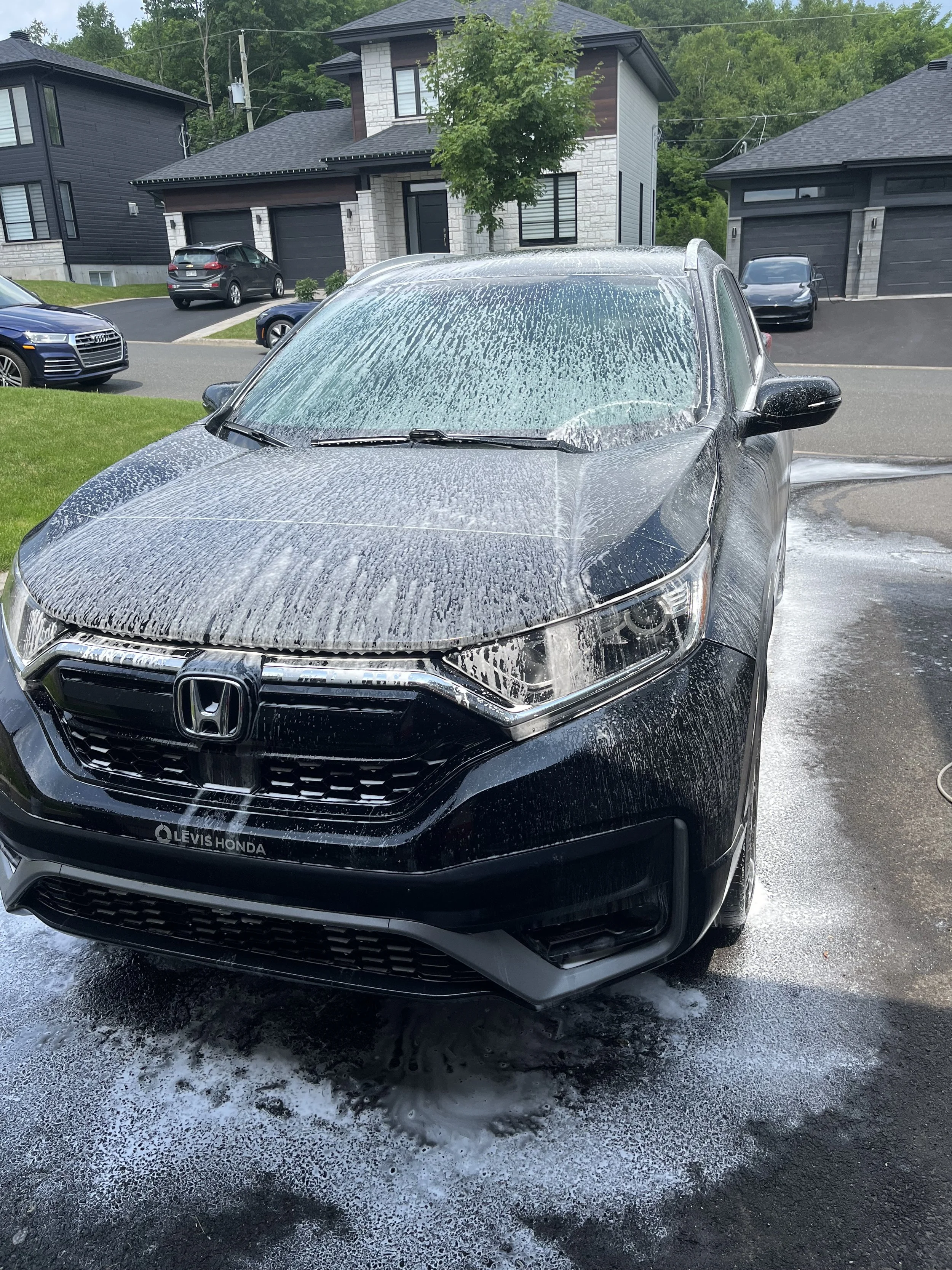 Voiture noire en cours de lavage avec du savon mousseux sur le capot et le pare-brise, dans une allée résidentielle.