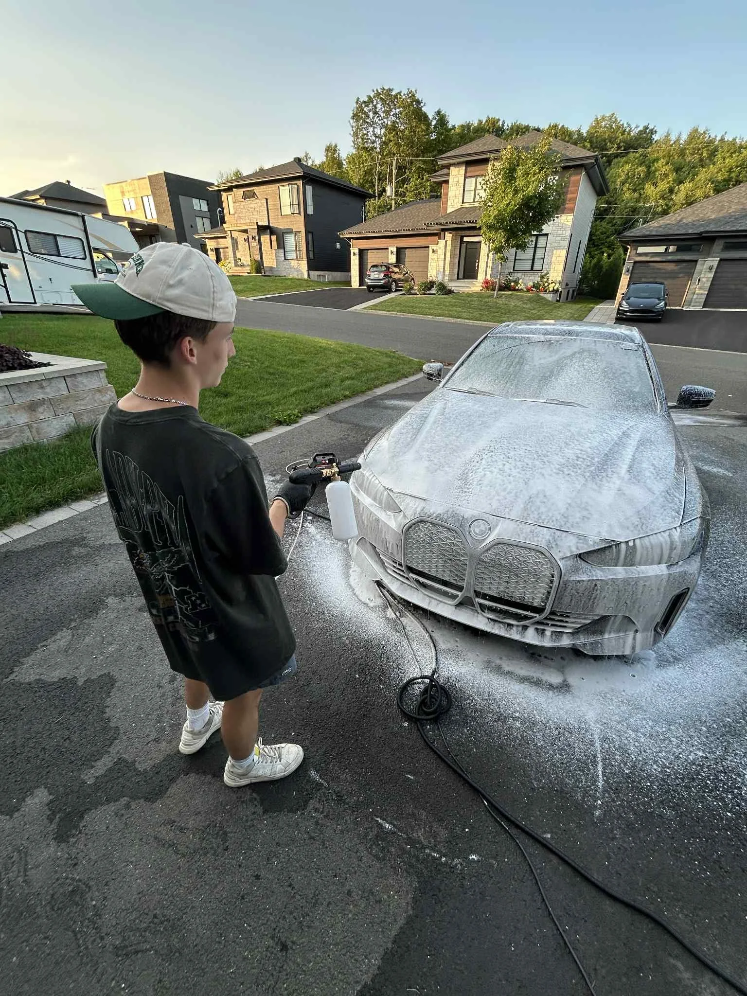 Un jeune garçon en t-shirt noir, portant une casquette et des gants, lave une voiture grise avec de la mousse dans une allée résidentielle. La scène se déroule en plein jour avec des maisons en arrière-plan et un ciel clair.