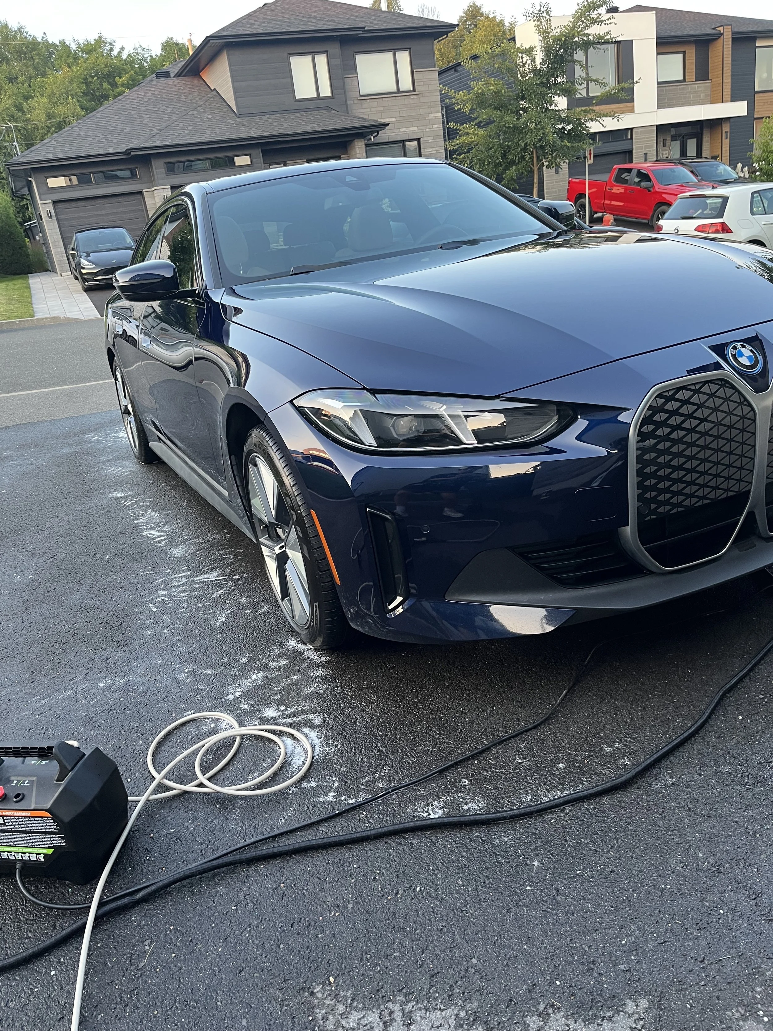Une voiture BMW noire en cours de nettoyage à domicile, avec un appareil de nettoyage et des câbles sur le sol asphalté.