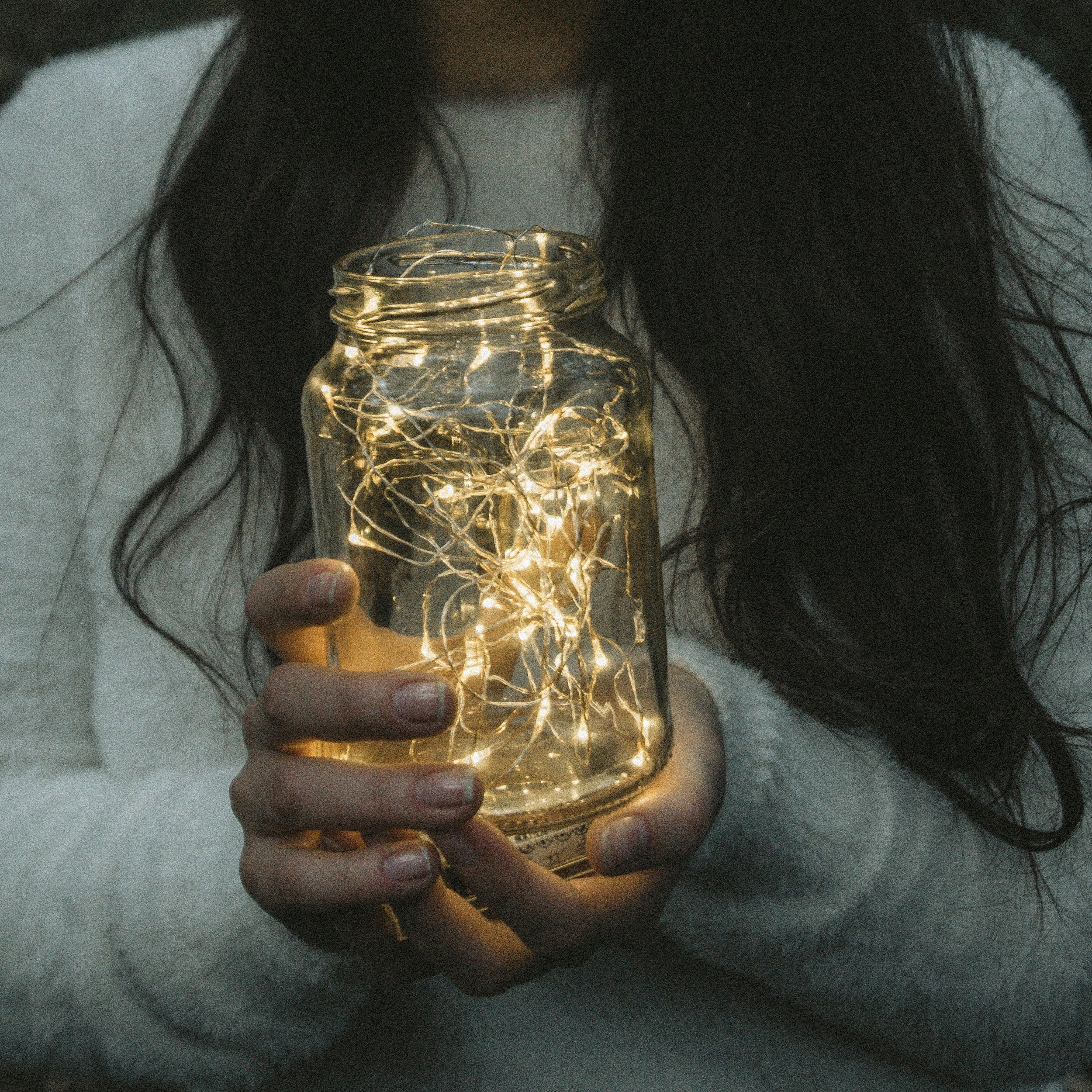 someone with long dark hear, wearing white sweater holds out a jar of twinkle lights to the camera.