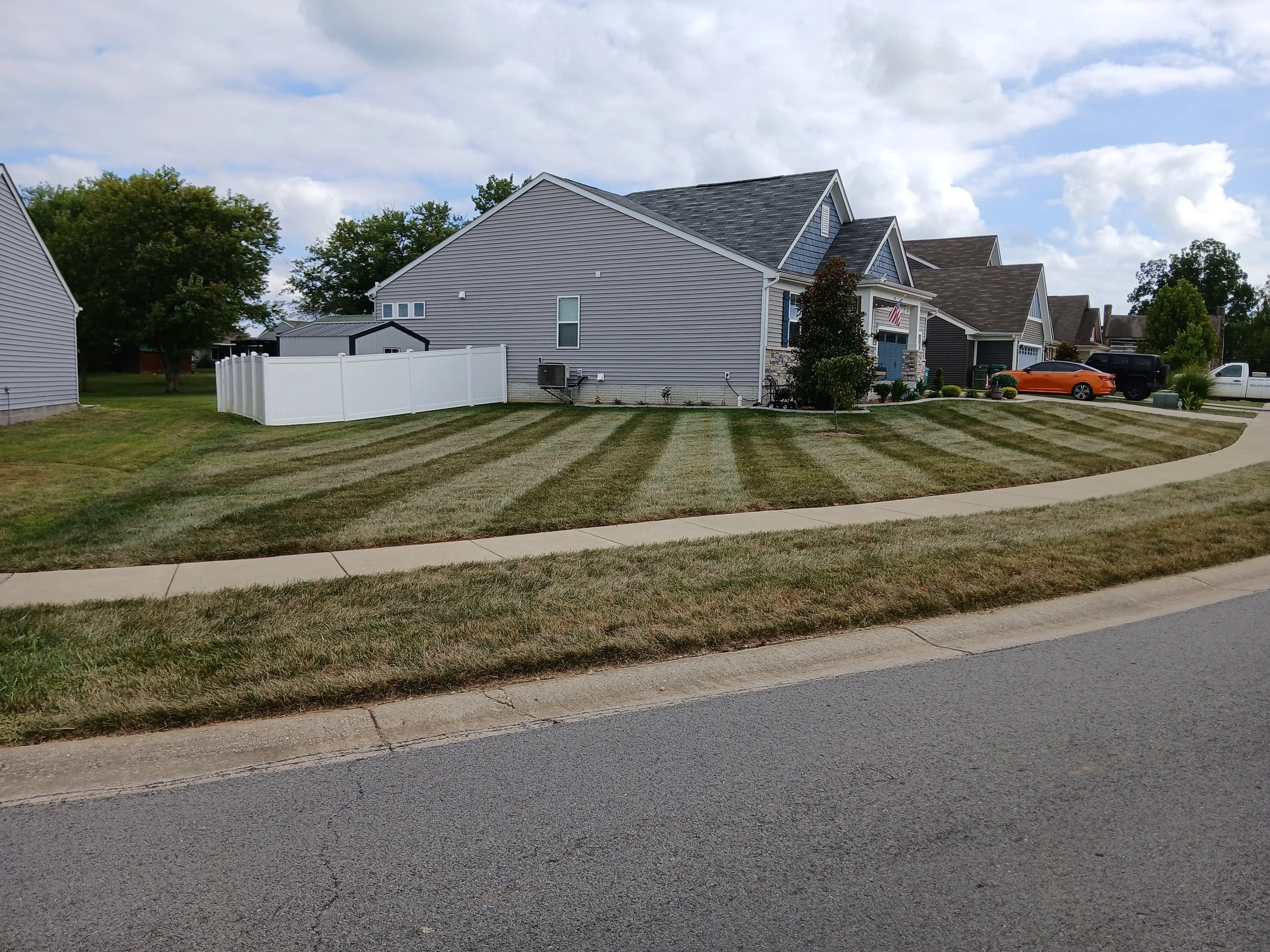 Residential neighborhood with a well-manicured front yard, sidewalk, and houses with cars parked in driveways. The sky is partly cloudy.