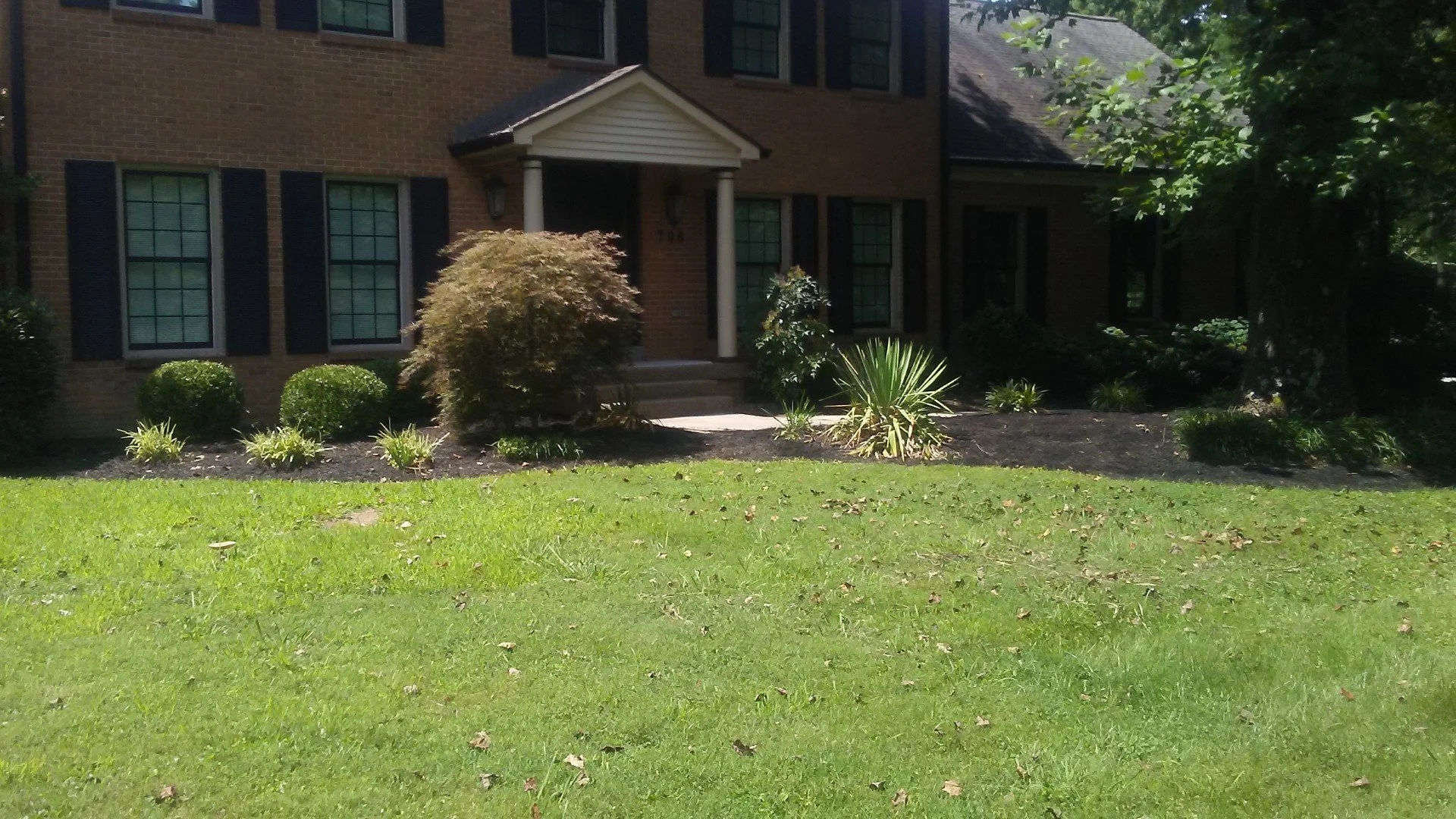 Front view of a brick house with four large windows having black shutters, a small porch with white columns, and a well-maintained front yard with green grass, bushes, and small plants.