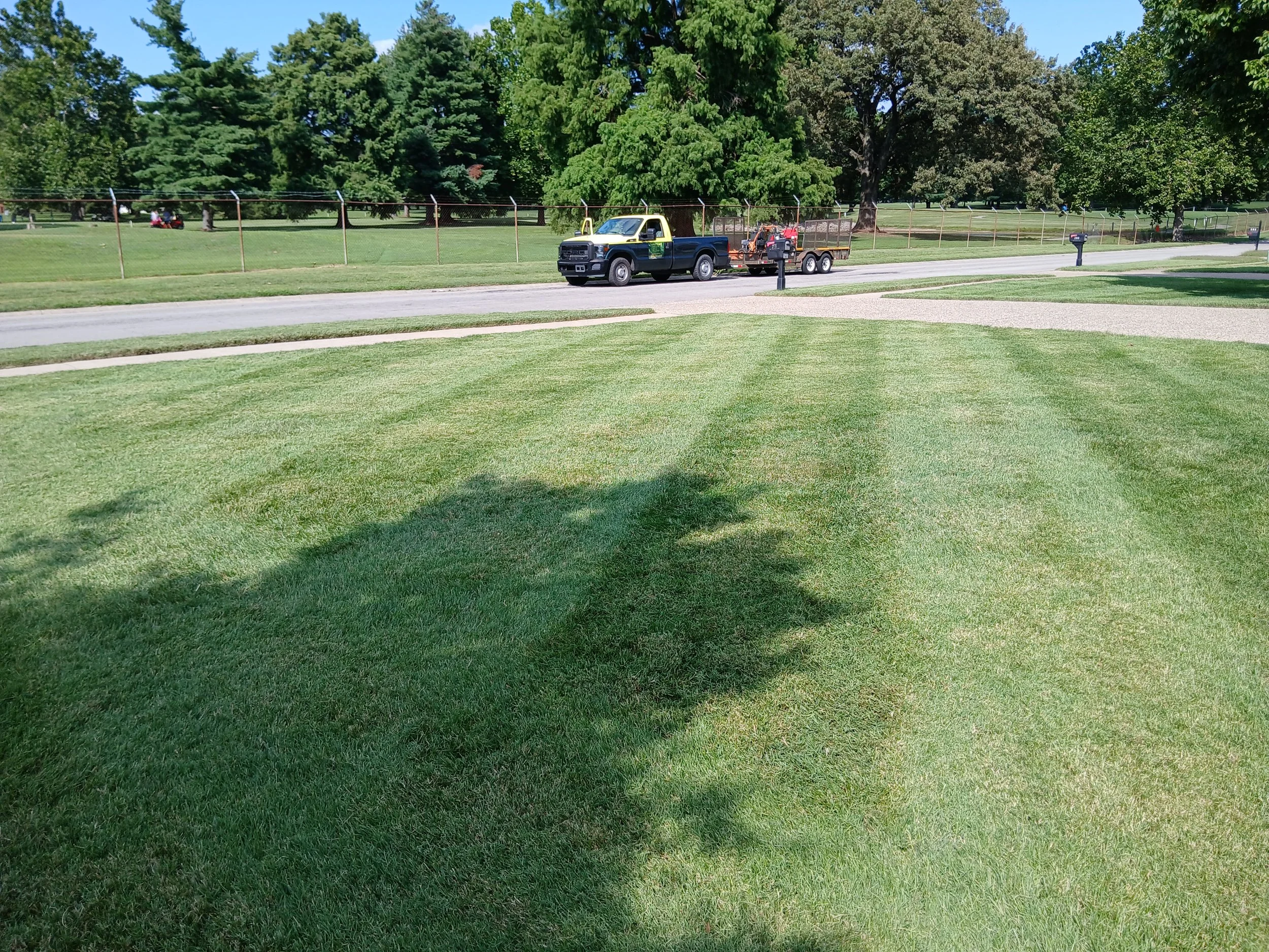 A yard with well-maintained grass and a large shadow from a tree, a paved walkway, a road, and a green and black pickup truck with a trailer parked alongside it, with trees and a fence in the background.