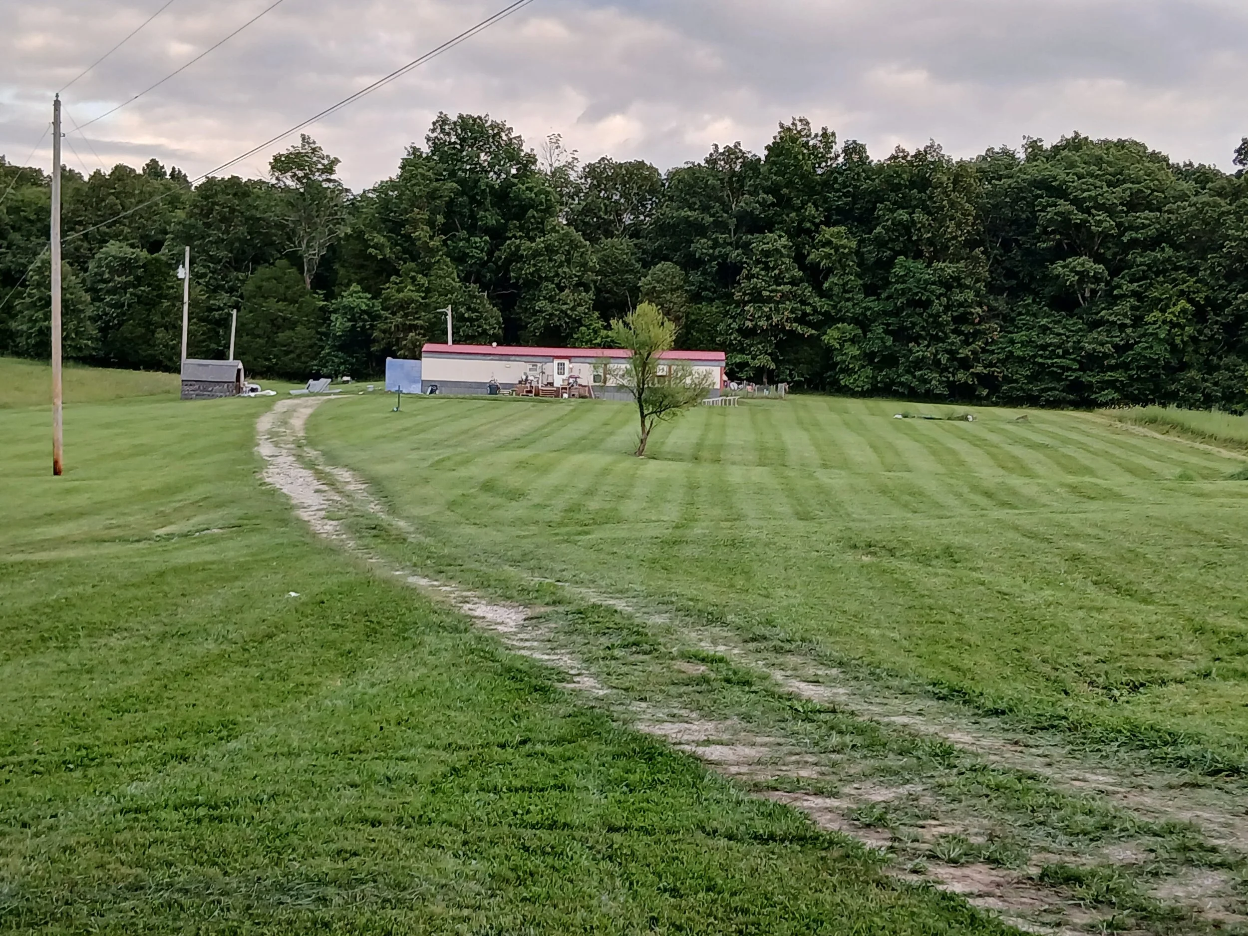 A grassy field with tire tracks leading toward a small house or mobile home, surrounded by trees and an overcast sky.